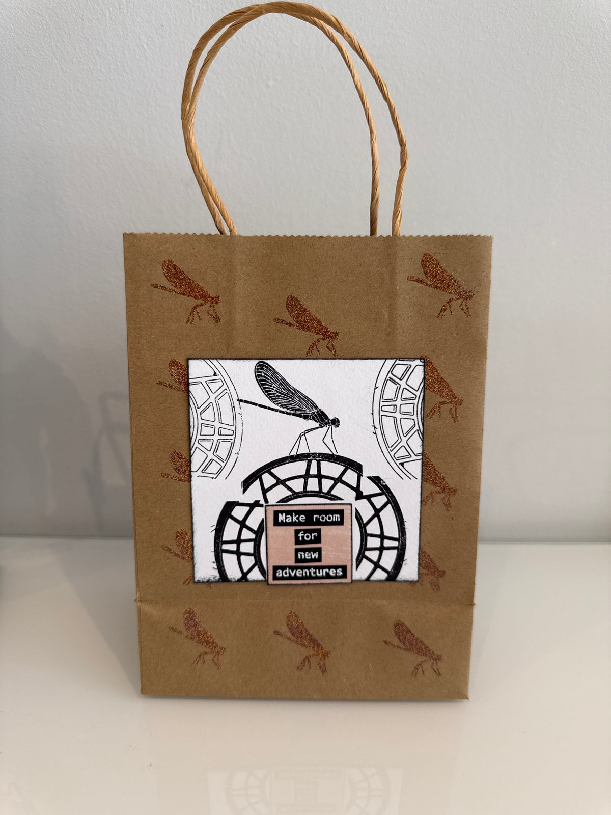 A brown kraft paper gift bag with twine handles, decorated with brown dragonfly illustrations and a central white label featuring a black dragonfly and the text 'MAKE ROOM FOR ADVENTURES'.