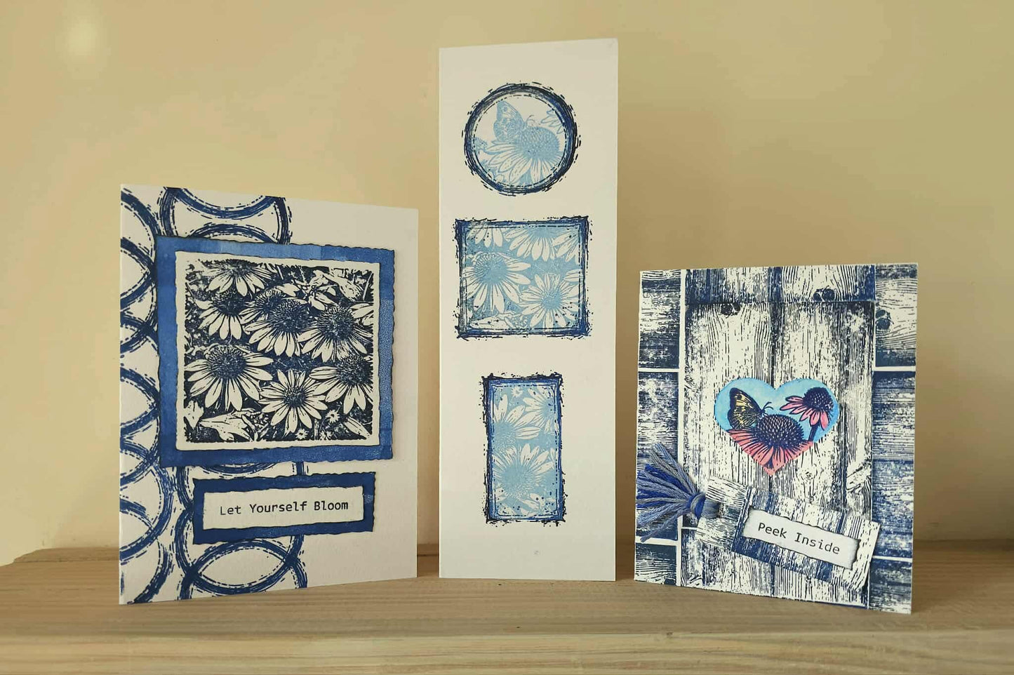 Three handmade greeting cards with blue and white floral and butterfly designs, displayed on a wooden surface against a beige background.