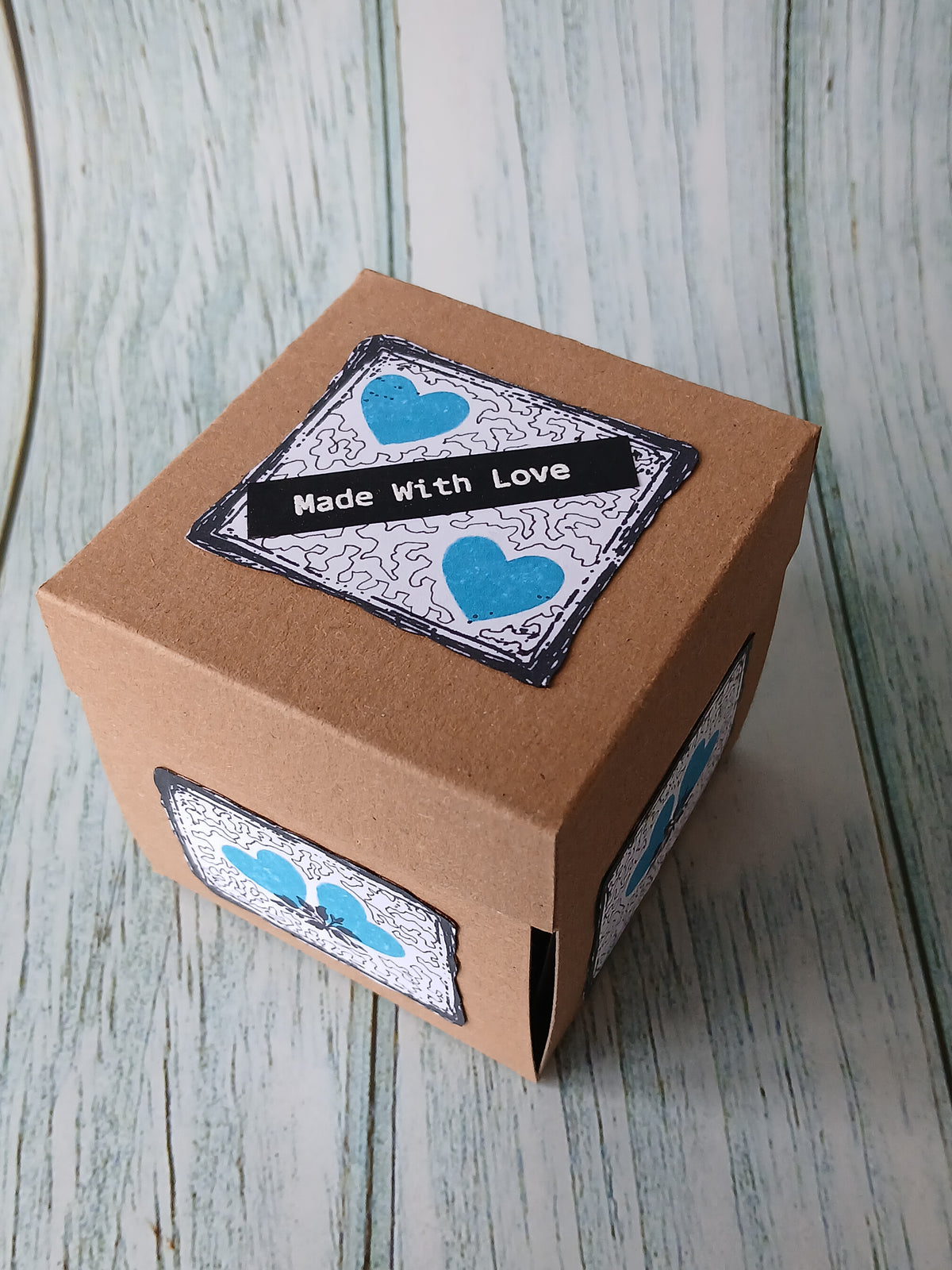 A handmade square gift box made of brown kraft paper, decorated with three decorative labels featuring blue hearts and the text 'Made with Love', set on a rustic light blue-gray wooden background.
