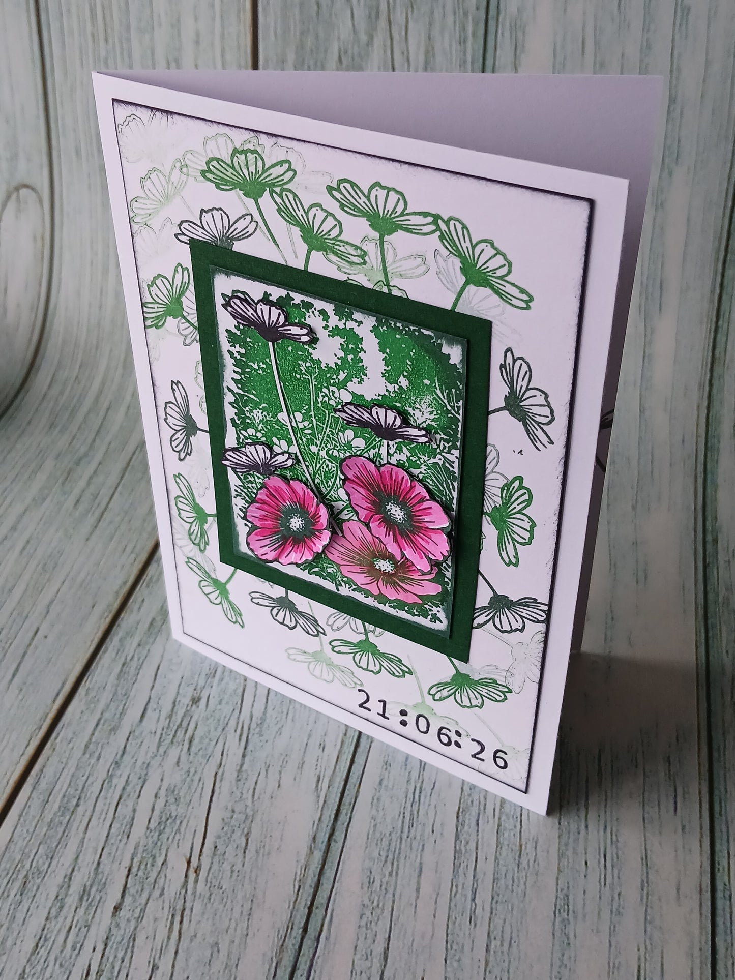 A handmade greeting card featuring a layered design with green and pink flowers, black and white botanical line art, and a digital clock timestamp at the bottom, displayed on a rustic gray wooden surface.