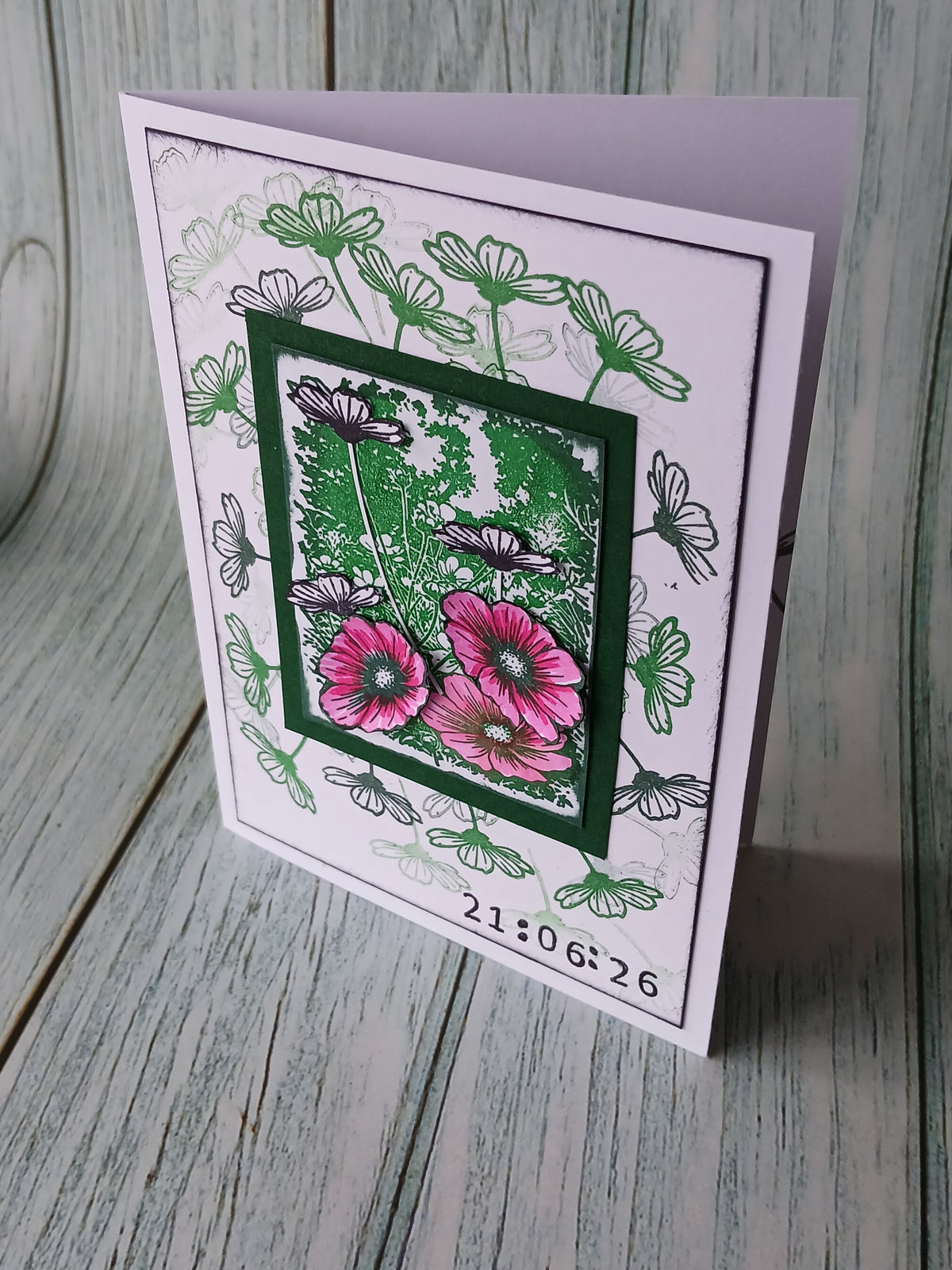 A handmade greeting card featuring a layered design with green and pink flowers, black and white botanical line art, and a digital clock timestamp at the bottom, displayed on a rustic gray wooden surface.