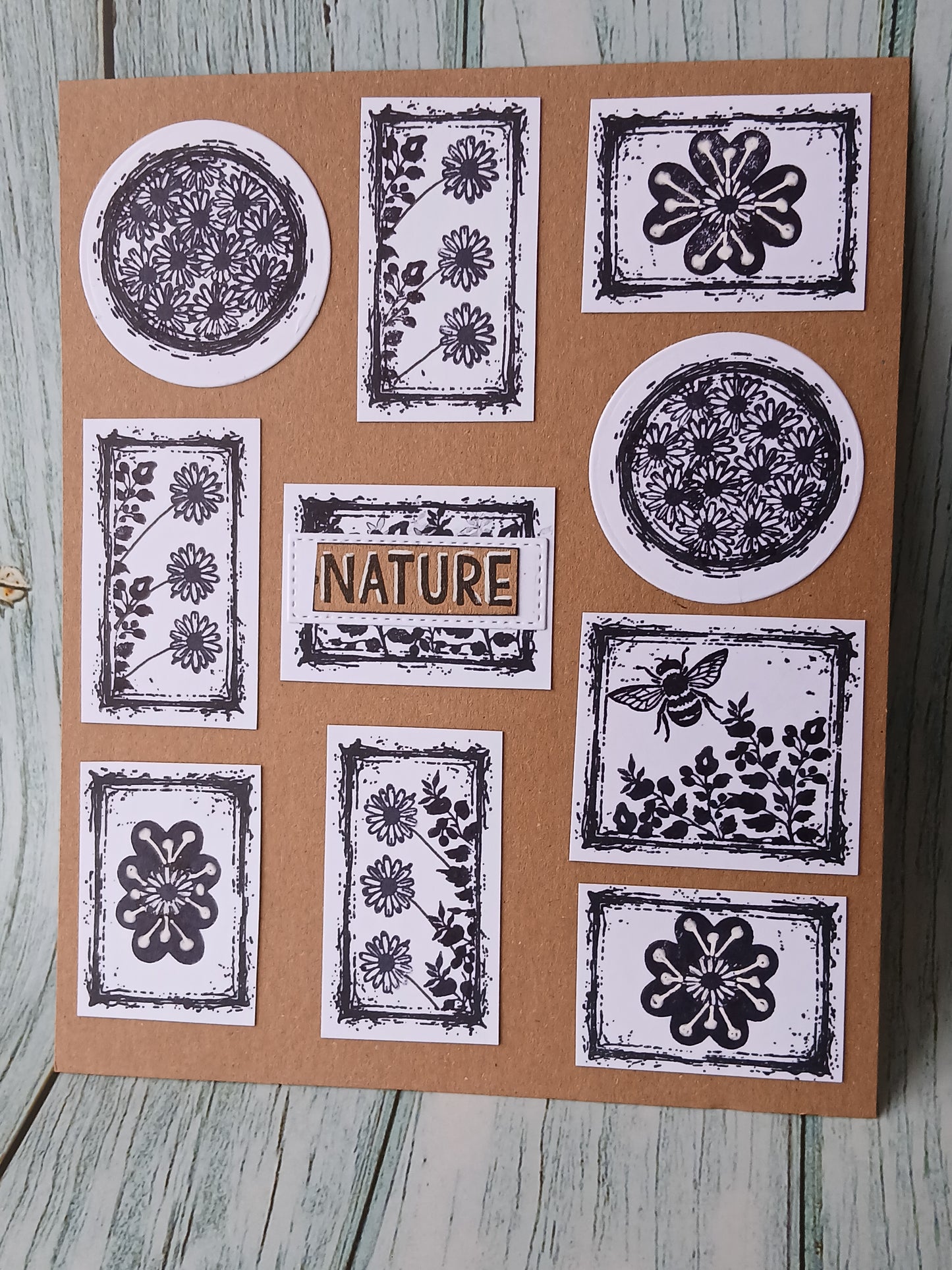 A square kraft paper card featuring multiple botanical and nature-themed stamp designs in black ink on white circular and rectangular stamps, with the word "NATURE" centered. The card is photographed standing on a distressed light blue-gray wooden surface.