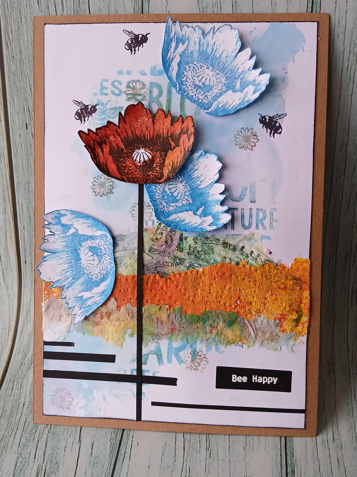 A handmade layered card featuring textured poppy flowers in blue, orange, and red, along with black bees and a watercolor landscape. The card is displayed on a light gray wooden surface.