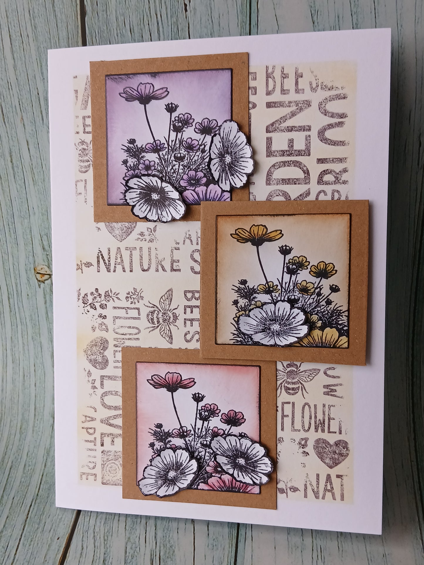 A handmade greeting card featuring three layered floral bouquets in purple, yellow, and pink, set against a background of vintage-style bee-themed text and illustrations, displayed on a rustic wooden surface.