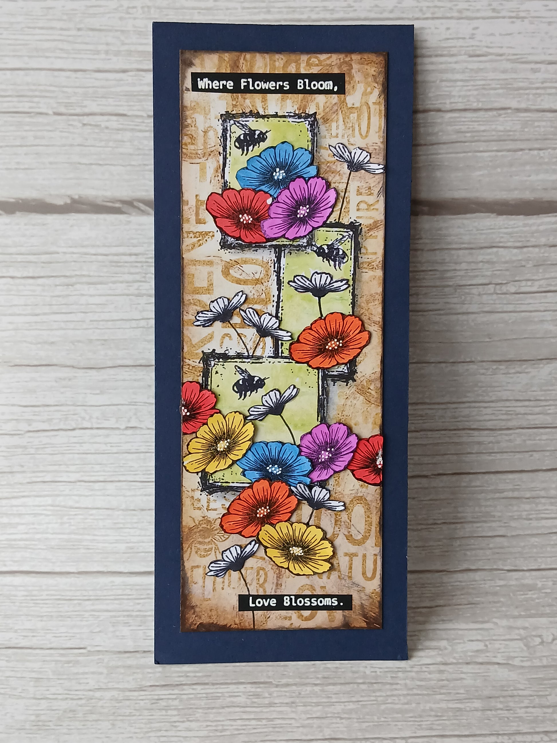 A vertical rectangular handmade card or plaque with a dark navy blue border, featuring colorful flowers, bees, and the text 'Where Flowers Bloom, Love Blossoms.' on a light wood background.