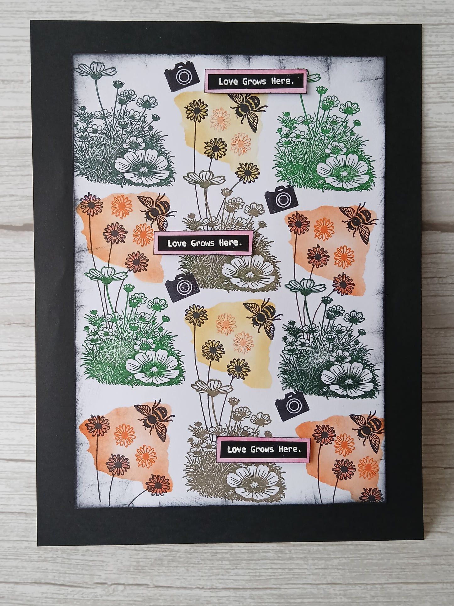 A framed art print featuring a retro-style collage of wildflowers, bees, and camera icons with the text 'Love Grows Here.