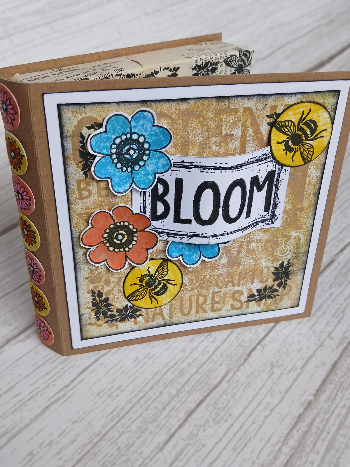 A handmade scrapbook album or journal with a kraft paper cover, featuring a decorated front cover with the word 'BLOOM' on a banner, colorful paper flowers, bee stickers, and floral accents, photographed on a light wood surface.