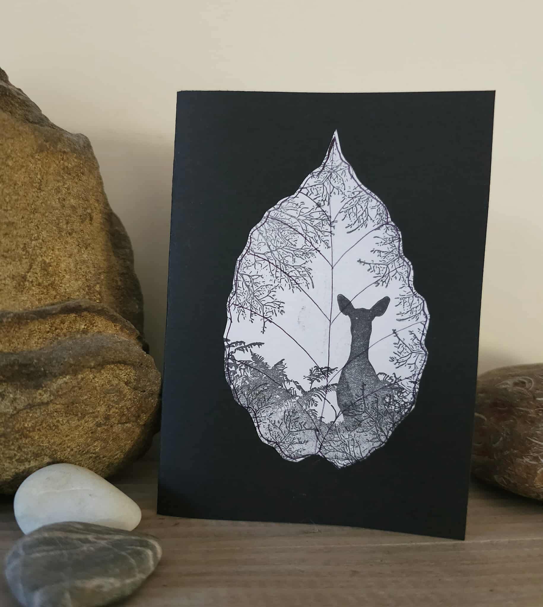 Black card with white leaf design featuring a deer silhouette, placed on a wooden surface with stones in the background.