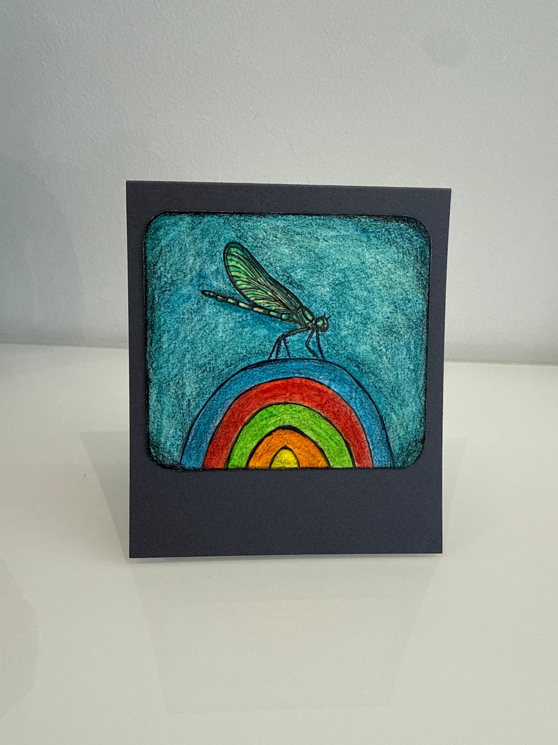 A square handmade art card featuring a colorful rainbow and dragonfly illustration on a dark navy blue cardstock, displayed on a white surface against a light gray background.