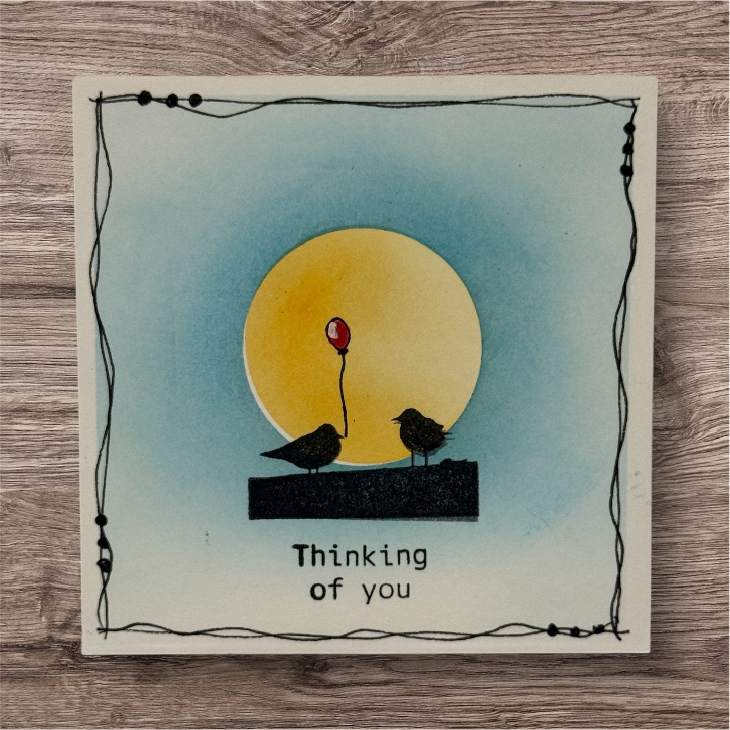 Card with two birds and a red balloon against a yellow sun, on a wooden surface.