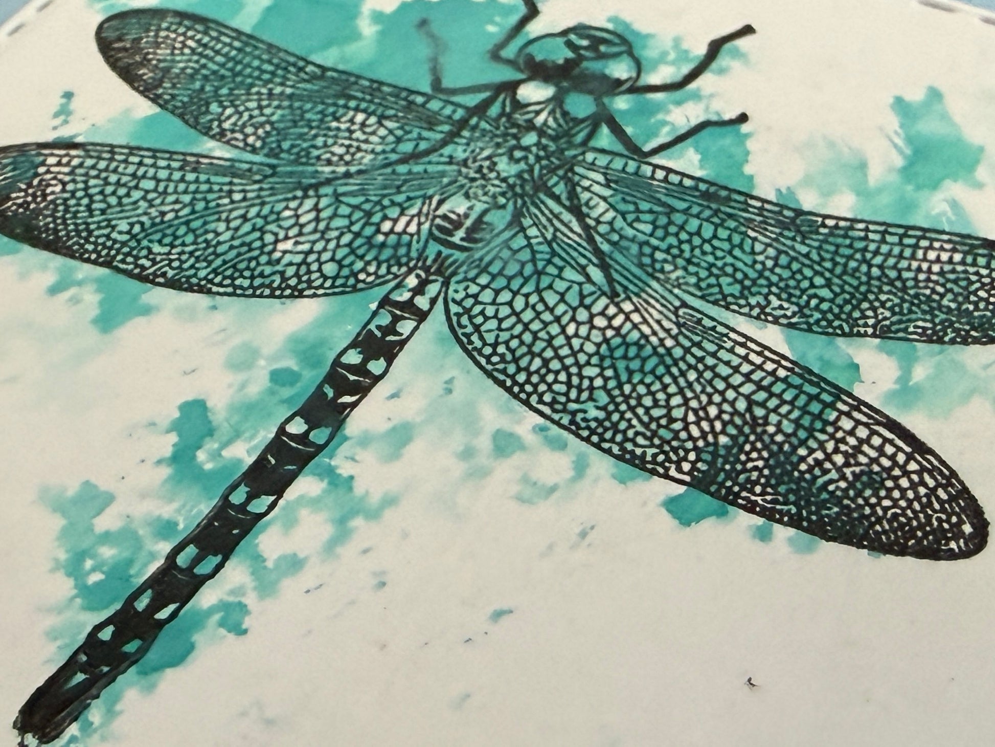 A close-up of a dragonfly artwork with a black ink-style body and wings over a white background with teal watercolor splashes.