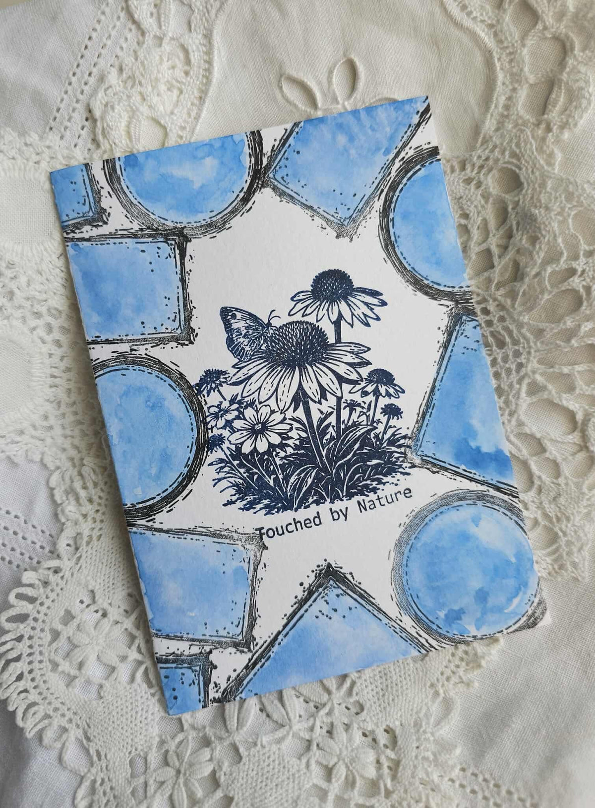 A square art print or notebook cover featuring a white background with blue watercolor geometric shapes and a black ink illustration of a butterfly and daisies. The text 'Touched by Nature' is printed near the bottom.
