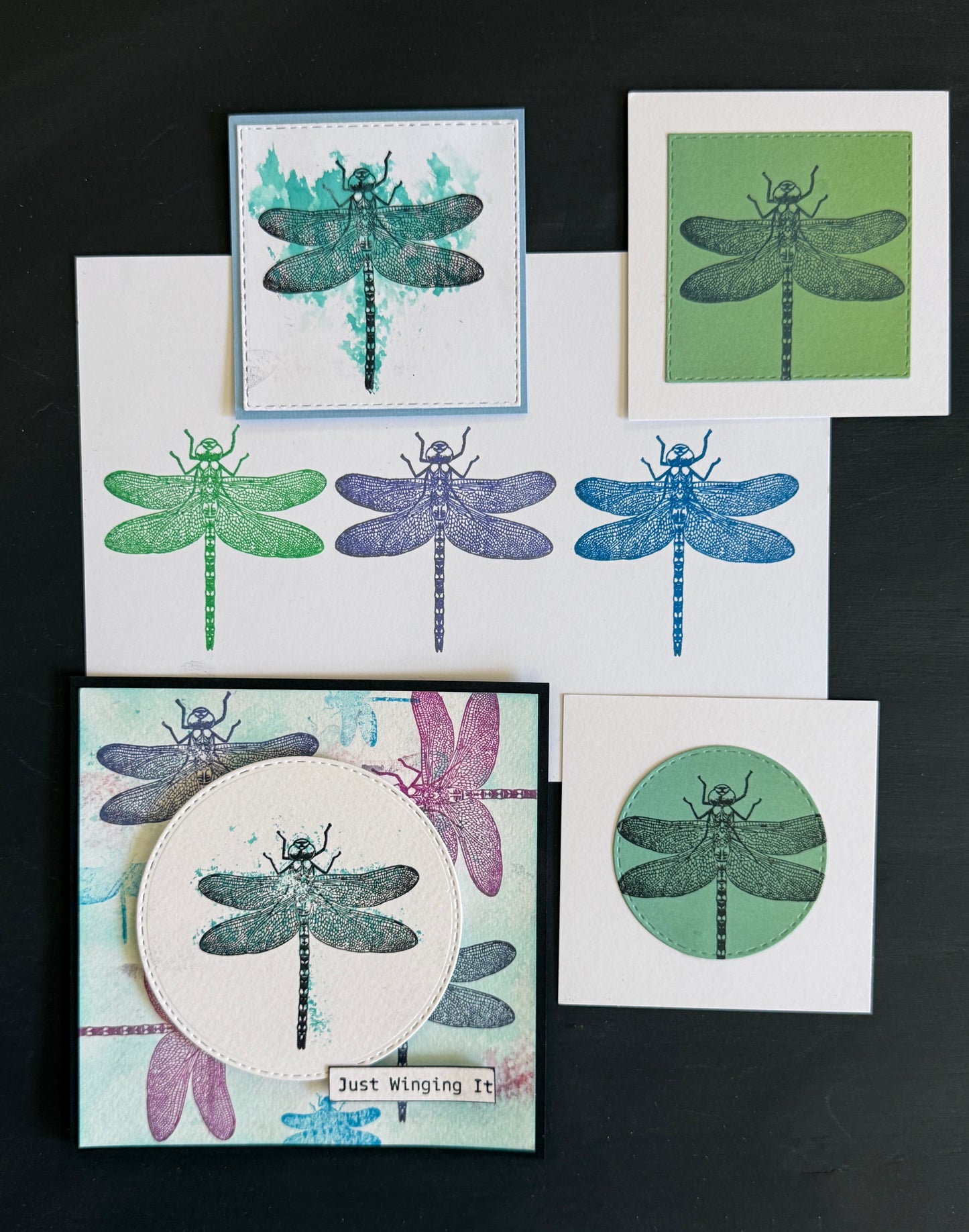 A set of handmade dragonfly-themed cards and stamp designs arranged on a dark background. The designs feature black dragonfly silhouettes with teal, green, blue, purple, and pink watercolor washes on white cardstock, including one card with a circular cutout and the phrase 'Just Winged It!'.