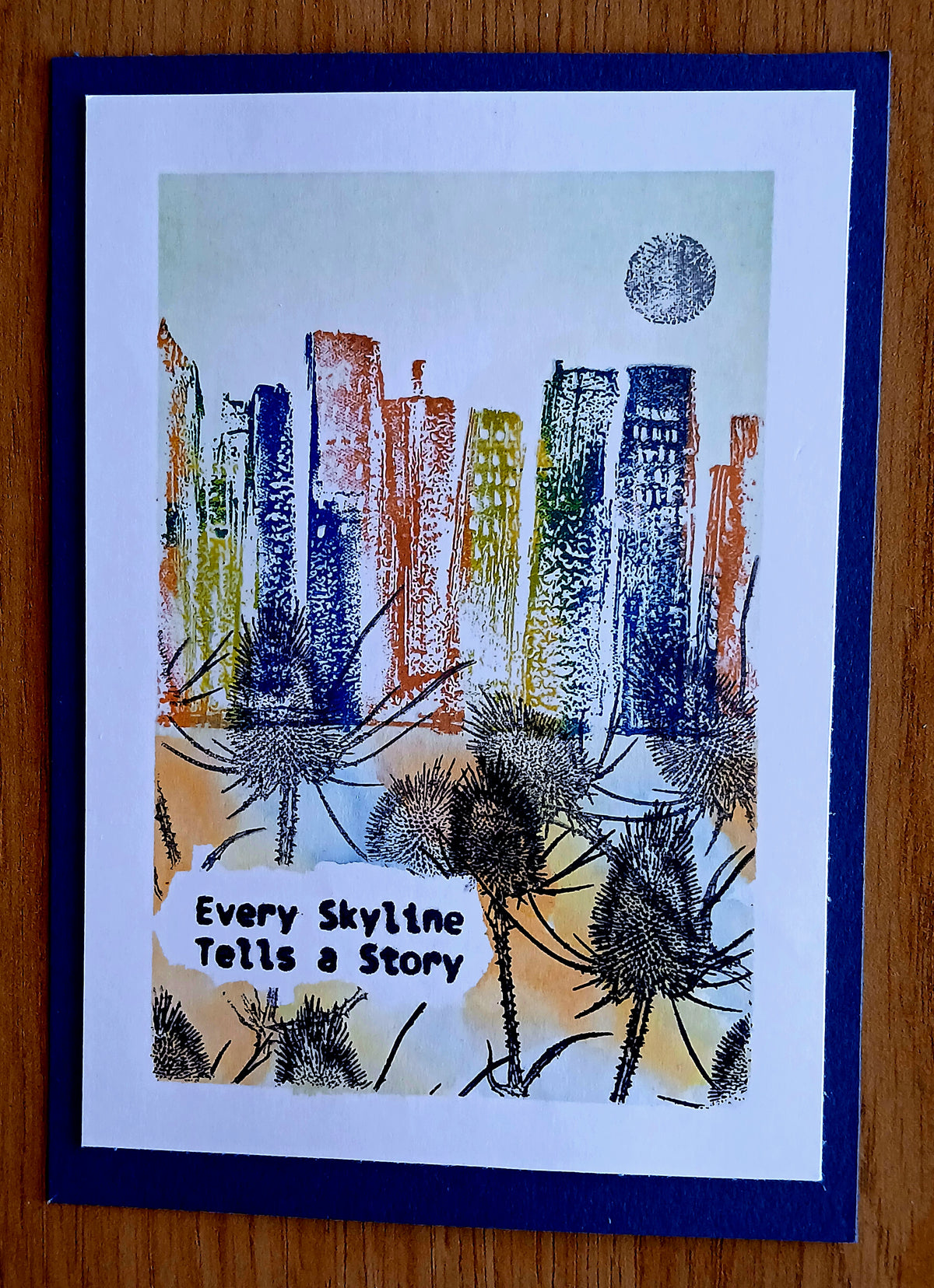 A square handmade art card featuring a colorful abstract skyline with skyscrapers, dandelions, and a moon, with the text 'Every Skyline Tells a Story' on a white background, bordered by a dark blue mat on a wooden surface.