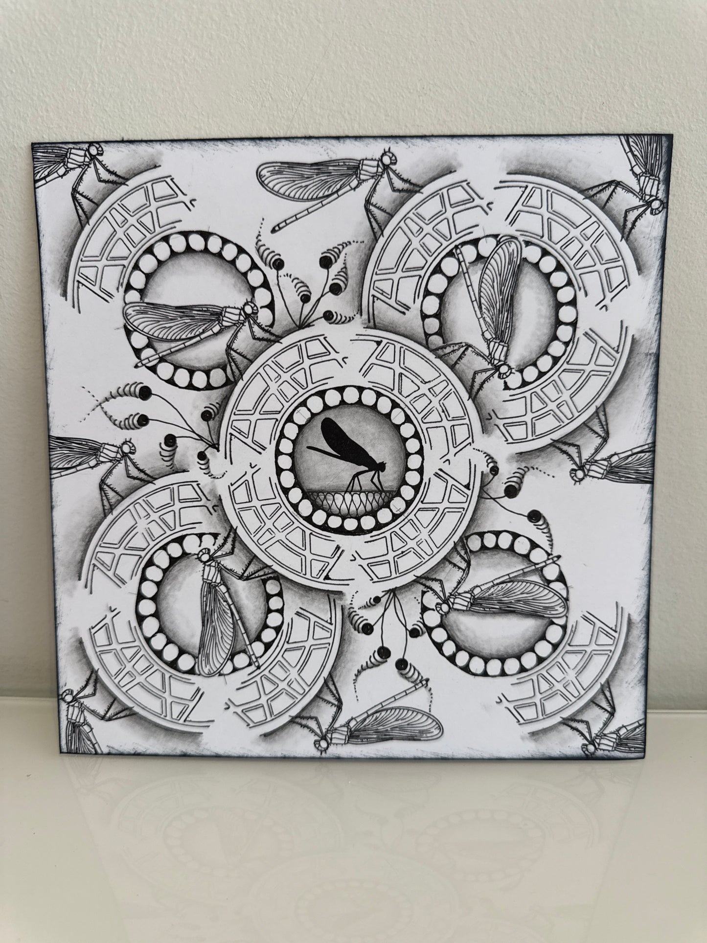 A square black and white art print or canvas featuring intricate insect and circular geometric patterns, photographed leaning against a light beige wall on a pale surface.