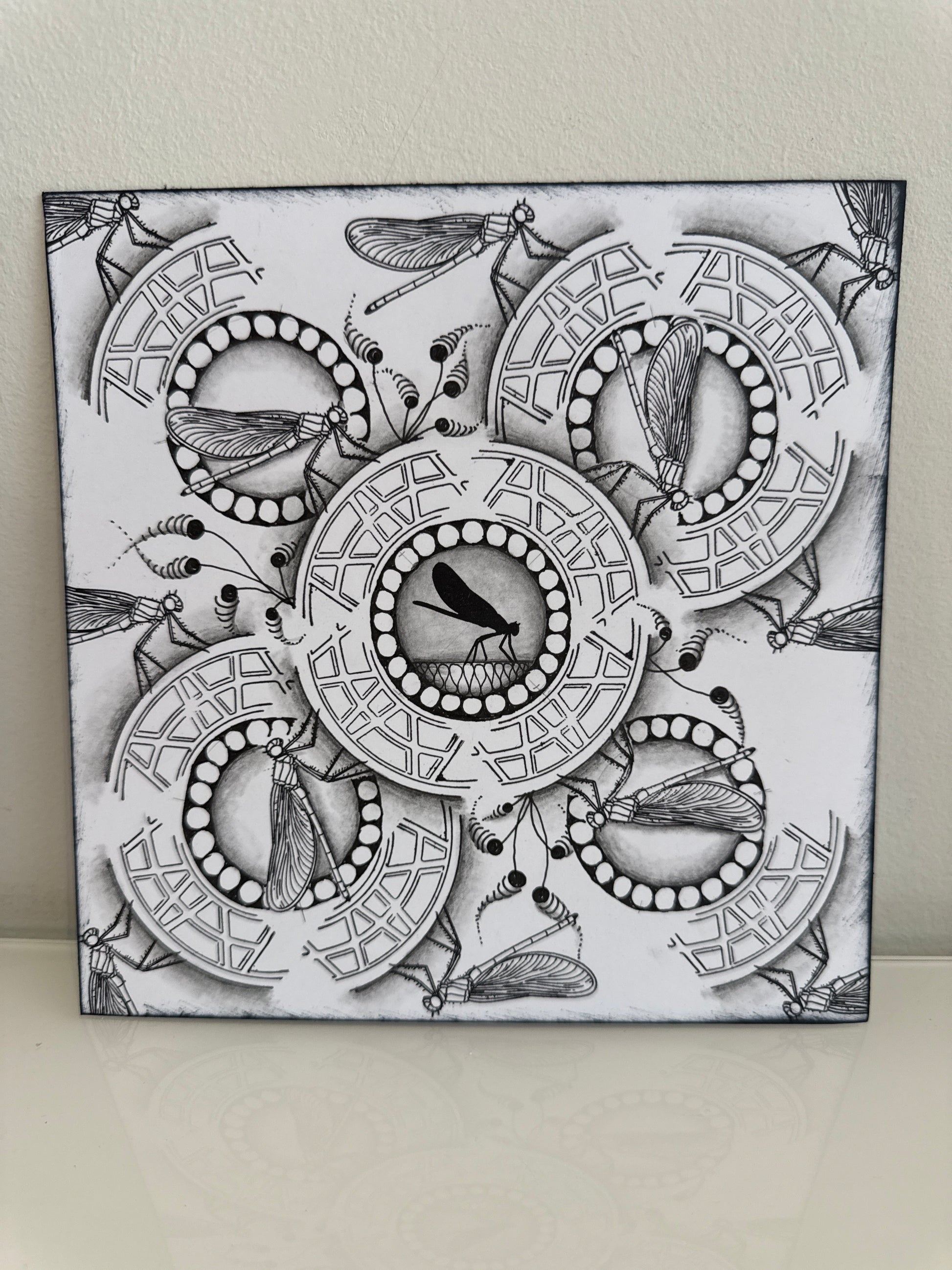 A square black and white art print or canvas featuring intricate insect and circular geometric patterns, photographed leaning against a light beige wall on a pale surface.