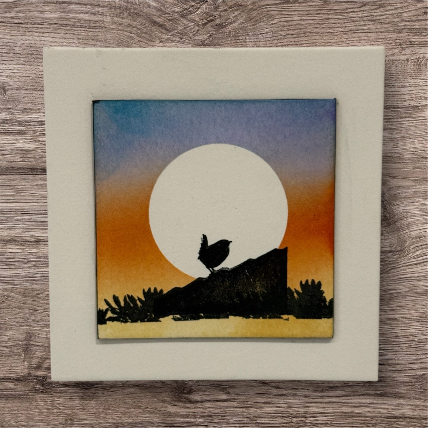 Framed artwork of a silhouette of a bird against a sunset sky on a wooden surface