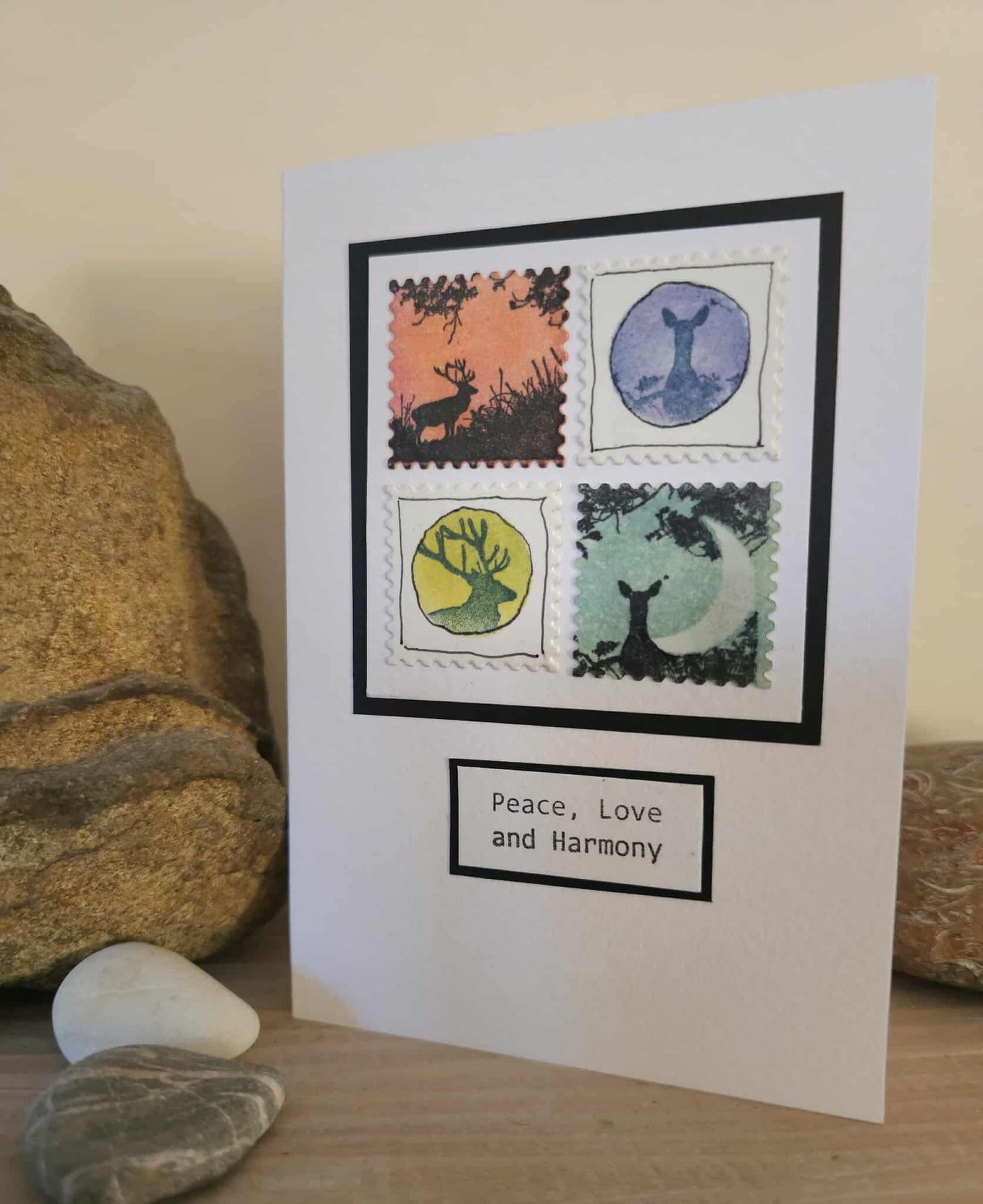 Card with stamp-like designs of deer and 'Peace, Love and Harmony' text, placed on a wooden surface with stones.