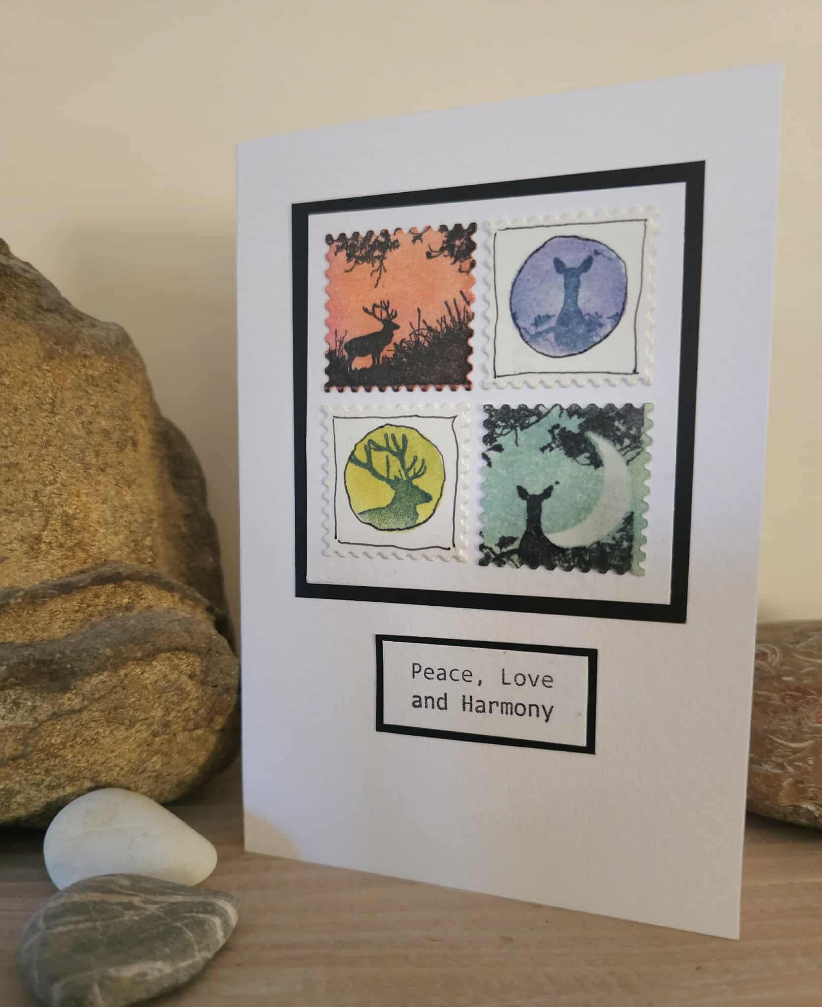 Card with stamp-like designs of deer and 'Peace, Love and Harmony' text, placed on a wooden surface with stones.