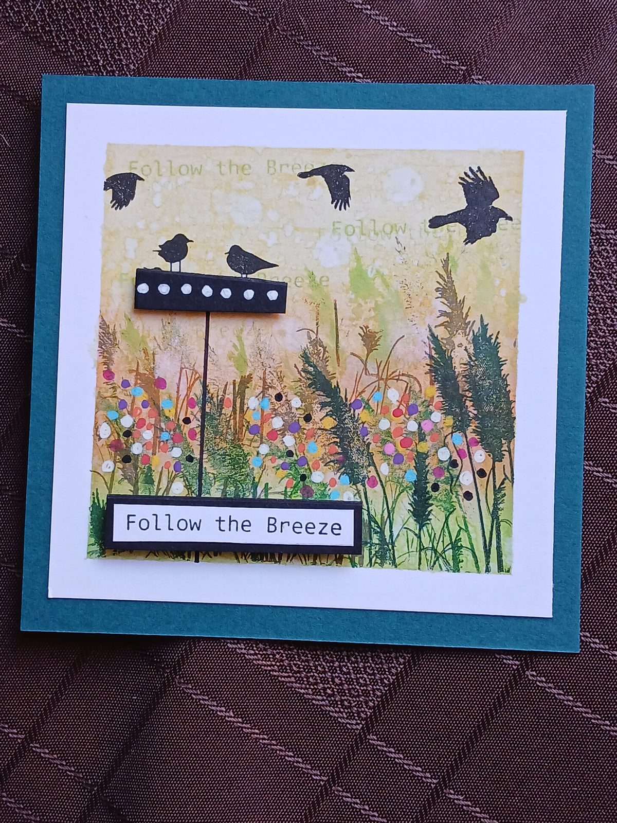 A handmade square greeting card featuring a bird feeder and birds over a colorful wildflower meadow, with the text 'Follow the Breeze' on a small black plaque. The card has a white inner border, a teal-blue outer border, and is photographed on a dark brown textured fabric background.