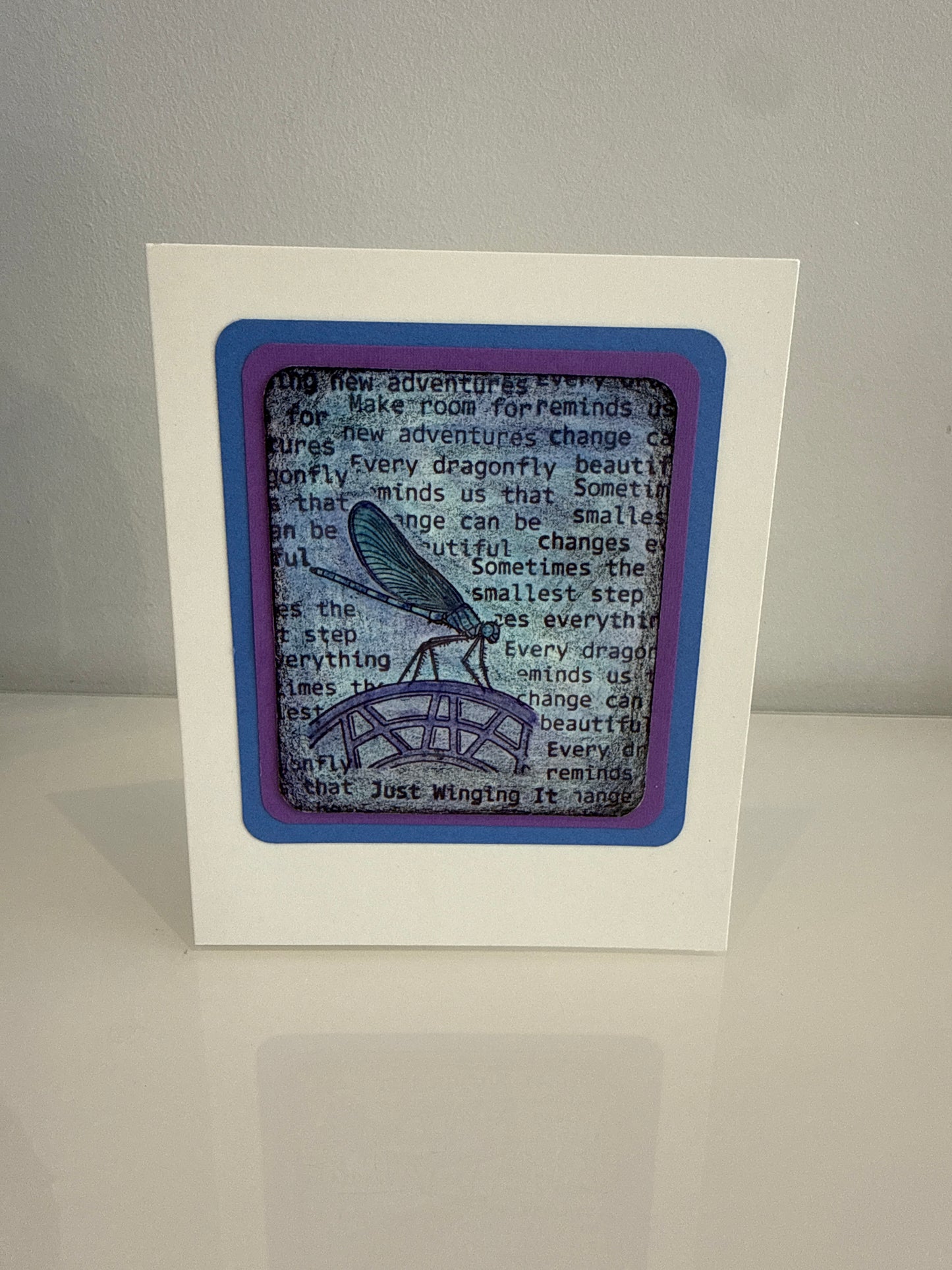 A square white card or framed print standing upright on a light surface against a light gray wall. The front features layered purple and blue borders surrounding a central printed design with black text and a blue winged figure on a gray background.