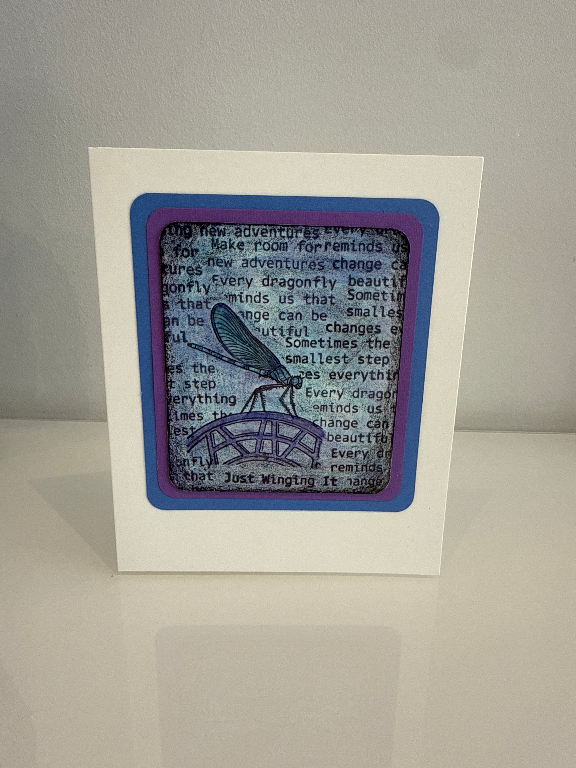 A square white card or framed print standing upright on a light surface against a light gray wall. The front features layered purple and blue borders surrounding a central printed design with black text and a blue winged figure on a gray background.