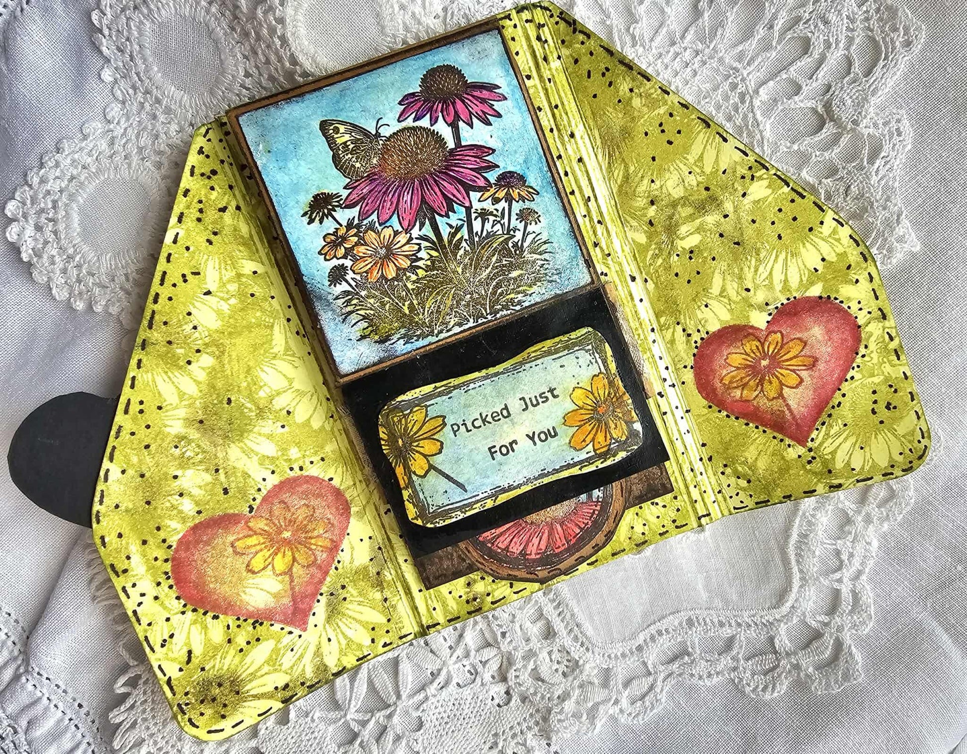 A handmade collage journal featuring a butterfly and coneflower design on the cover, decorated with yellow textured paper, black polka dots, and red heart-shaped flower accents, displayed on a white lace fabric background.