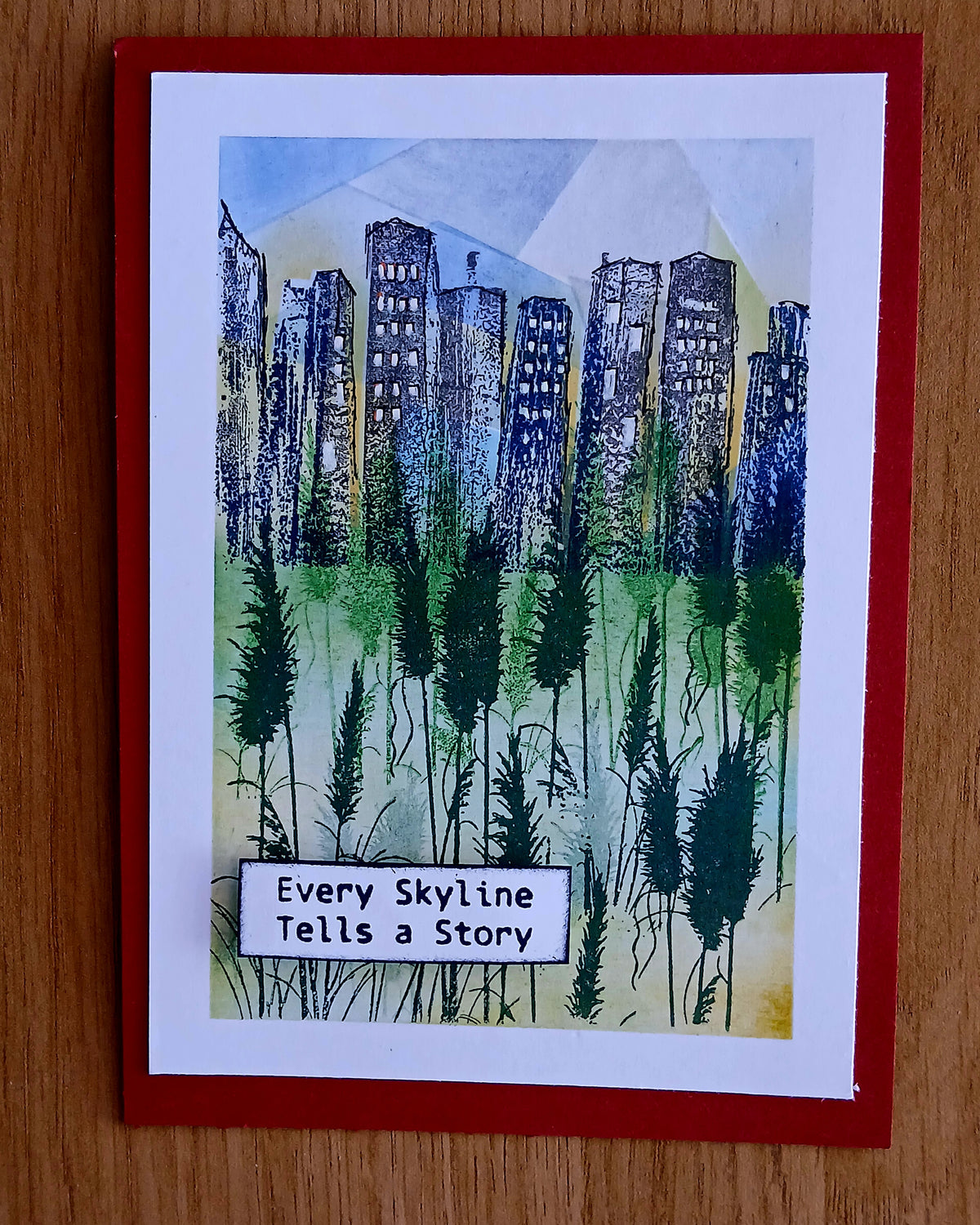 A square greeting card featuring a hand-painted style illustration of a city skyline with tall dark buildings and reeds in the foreground, with the text 'Every Skyline Tells a Story' at the bottom. The card has a white border and a red backing, shown on a wooden surface.