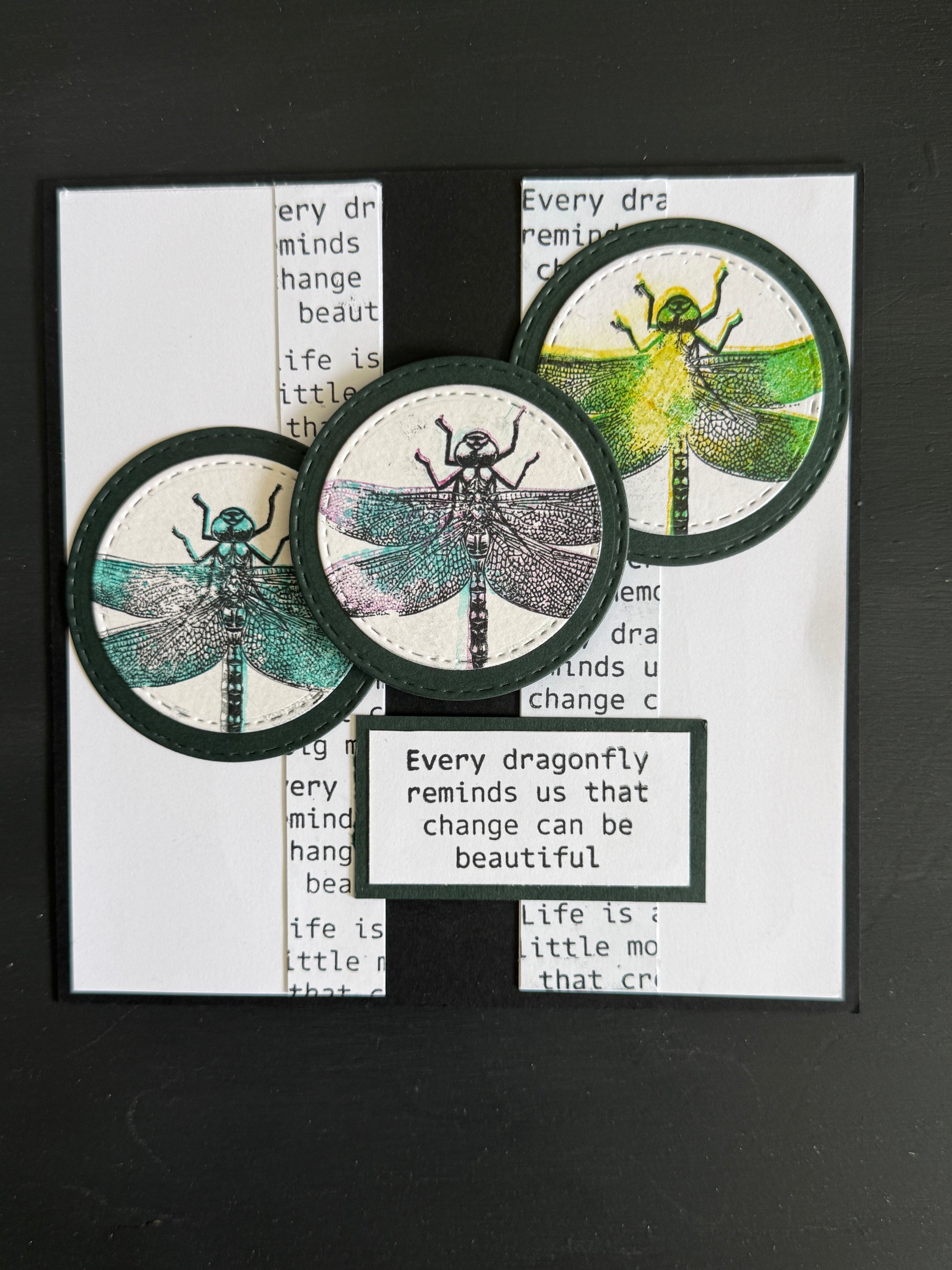 A handmade dragonfly-themed card featuring three circular dragonfly illustrations in teal, gray, and green, layered over a white card with printed text and a black banner that reads, 'Every dragonfly reminds us that change can be beautiful.'.