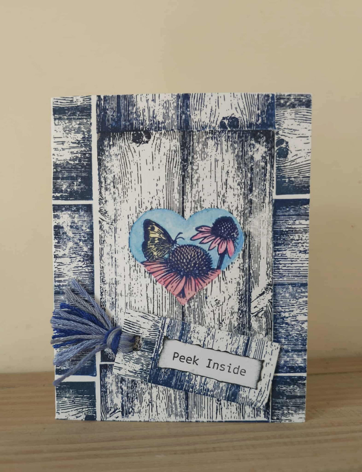 A handmade greeting card featuring a rustic blue and white weathered wood design, a heart-shaped butterfly and flower motif, a blue tassel, and a small tag that reads 'Peek Inside', displayed on a wooden surface against a beige background.