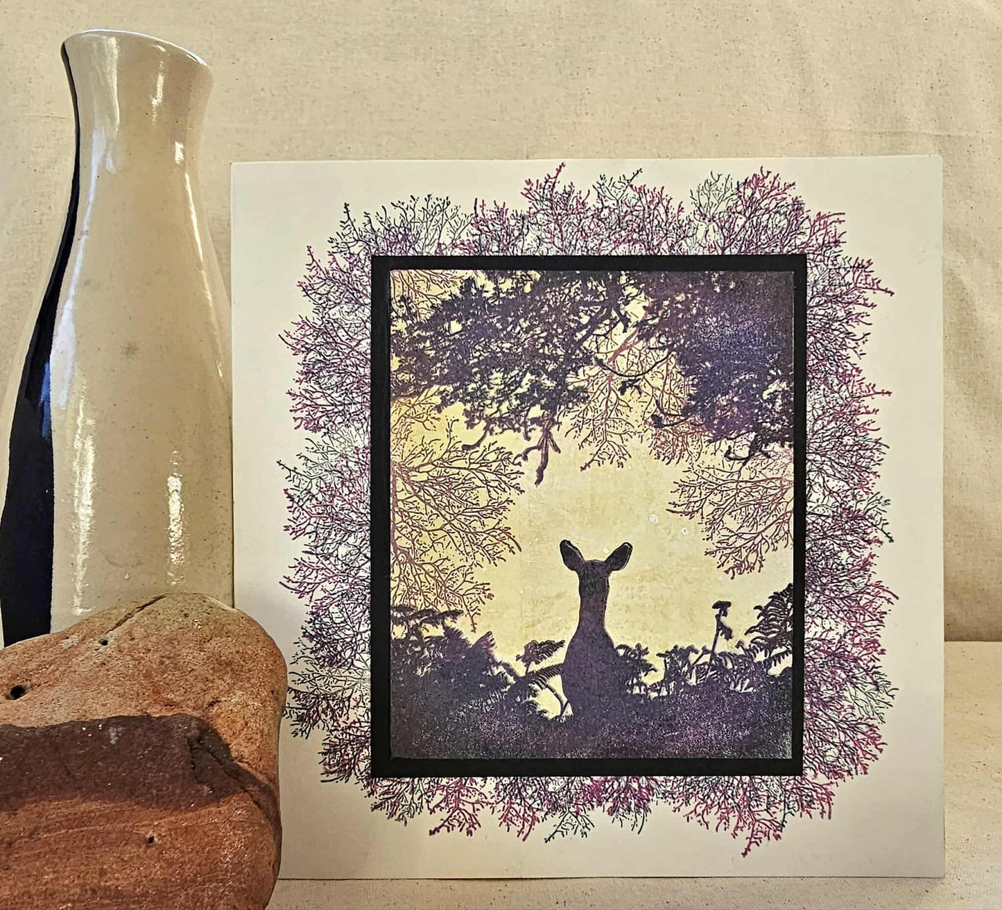 Framed artwork of a deer silhouette with a person in a forest setting, placed on a textured surface.