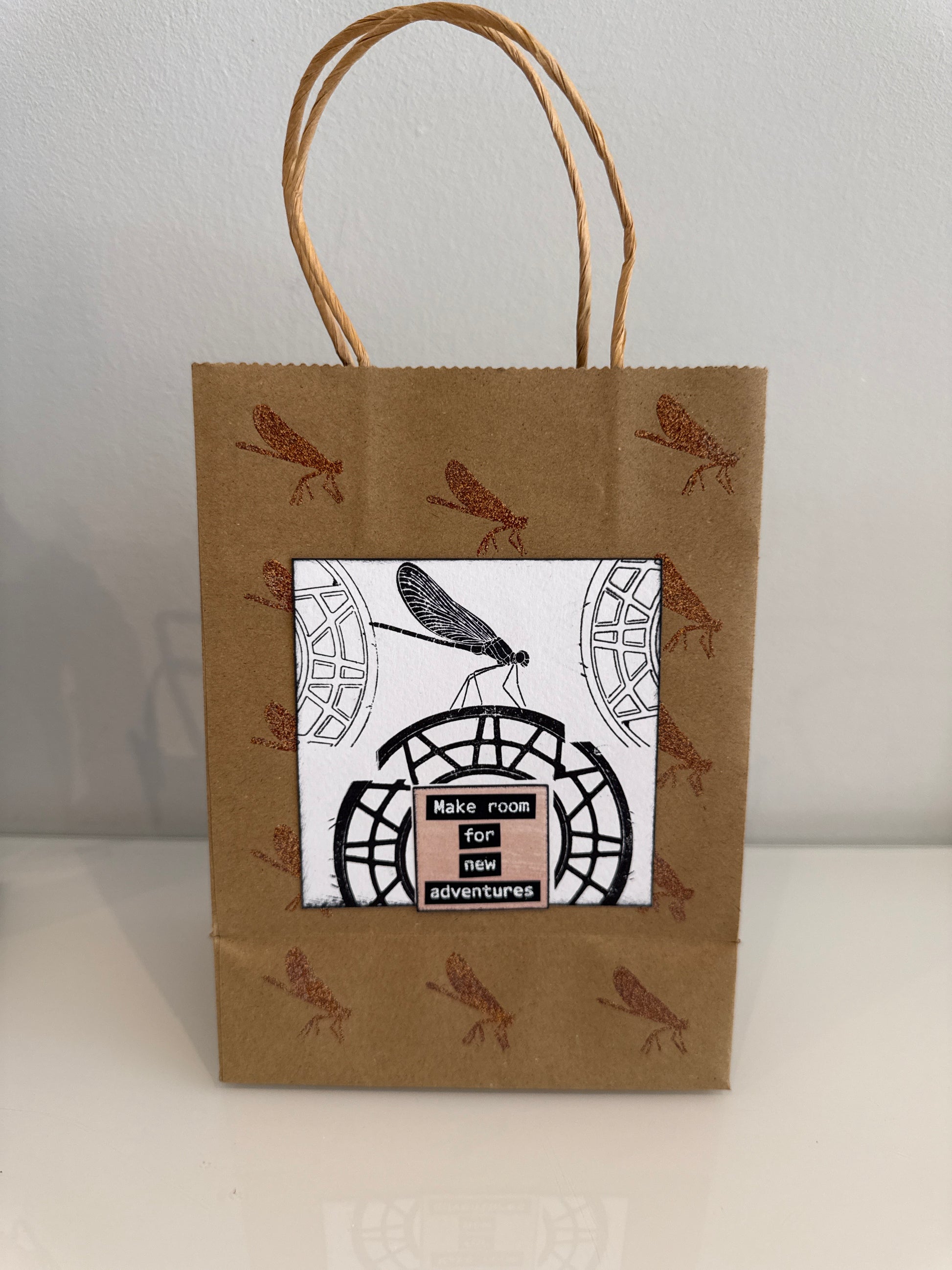 A brown kraft paper gift bag with twine handles, decorated with brown dragonfly illustrations and a central white label featuring a black dragonfly and the text 'MAKE ROOM FOR ADVENTURES'.