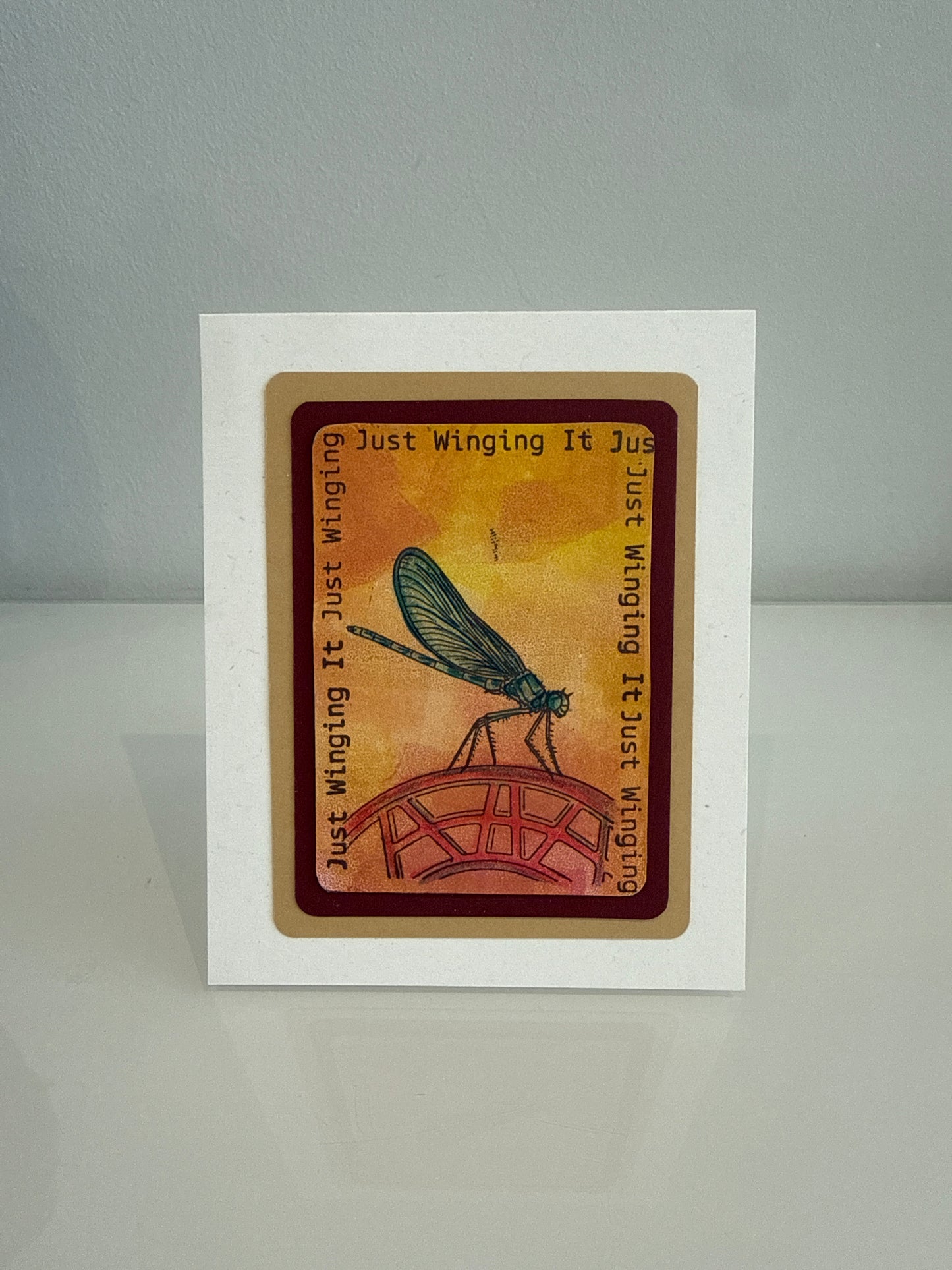 A square white card featuring a central printed illustration of a dragonfly on a red bridge against a warm orange-yellow background, with the phrase 'Just Winging It Just Winging' printed around the design. The card is photographed standing upright on a light gray surface against a gray background.