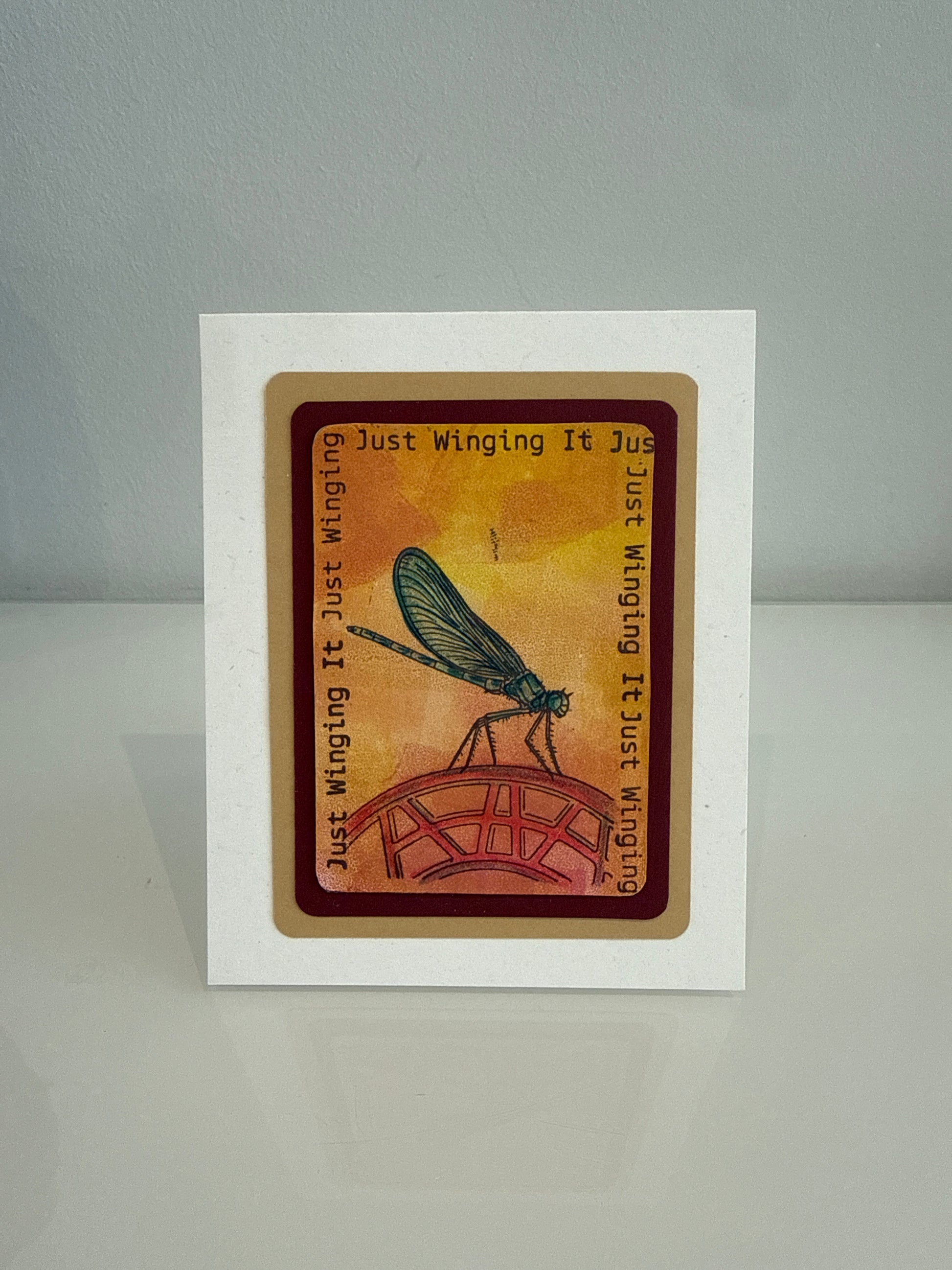 A square white card featuring a central printed illustration of a dragonfly on a red bridge against a warm orange-yellow background, with the phrase 'Just Winging It Just Winging' printed around the design. The card is photographed standing upright on a light gray surface against a gray background.