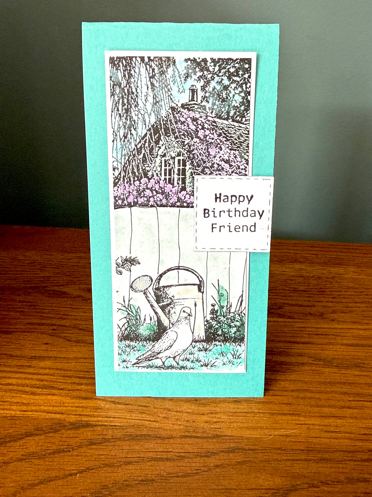 A handmade greeting card with a turquoise border and a black-and-white garden illustration featuring a house, flowers, a watering can, and a bird. A white label on the card reads 'Happy Birthday Friend.
