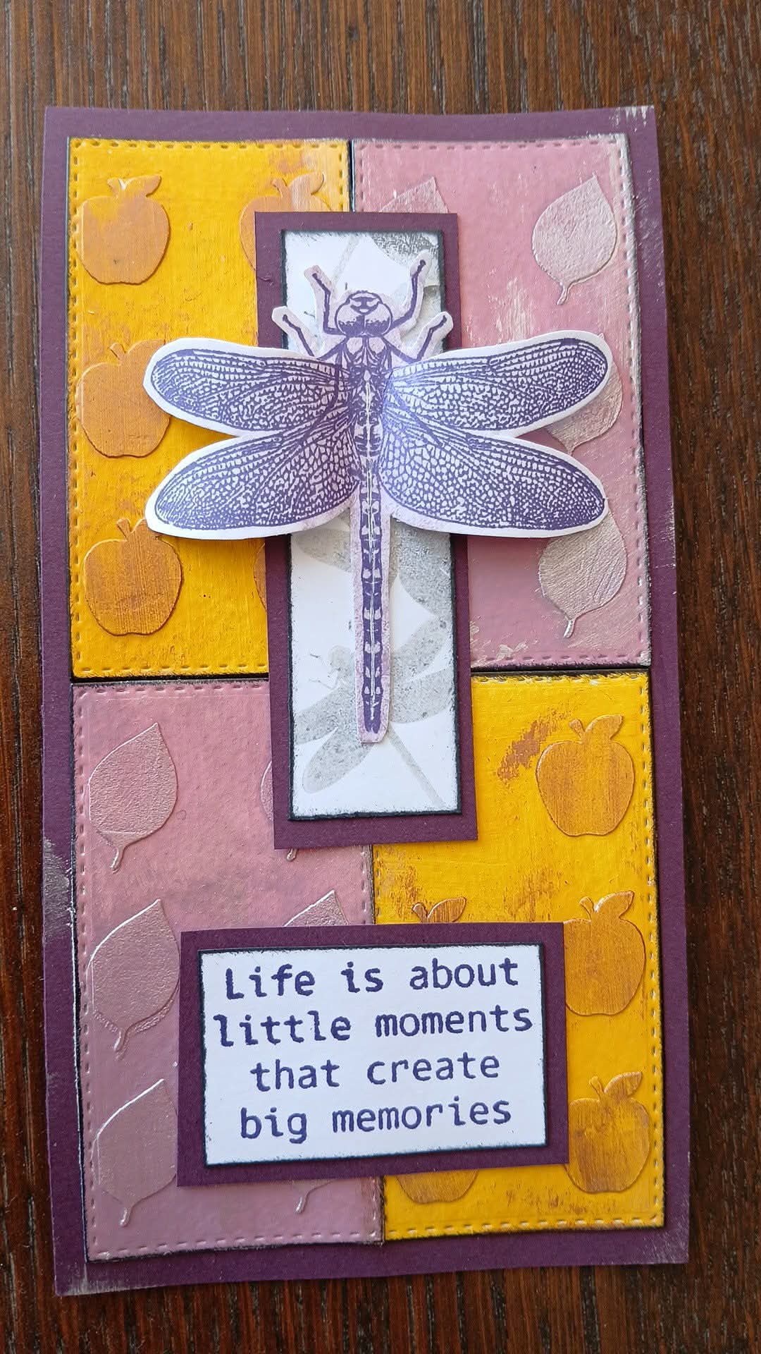 A handmade layered card featuring a die-cut dragonfly on a purple and white background, layered over yellow and dusty pink textured panels with embossed apple and leaf designs. A stitched border surrounds the card, and a white text box at the bottom reads, 'Life is about little moments that create big memories.