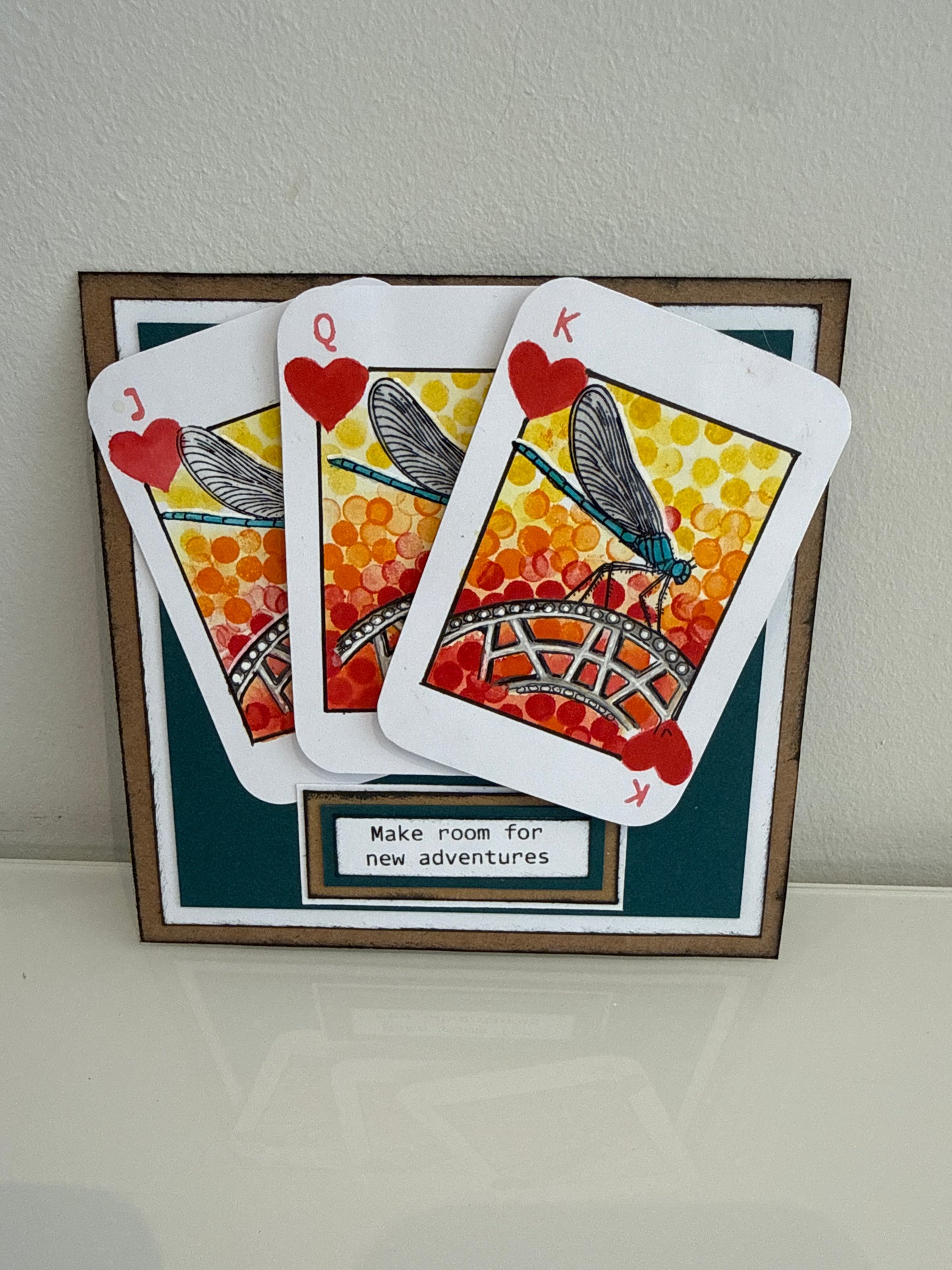 A framed decorative display featuring three overlapping playing cards styled like insect-themed cards with red hearts and orange-yellow patterns, mounted on a dark teal backing with a small label that reads 'Make room for new adventures.'.