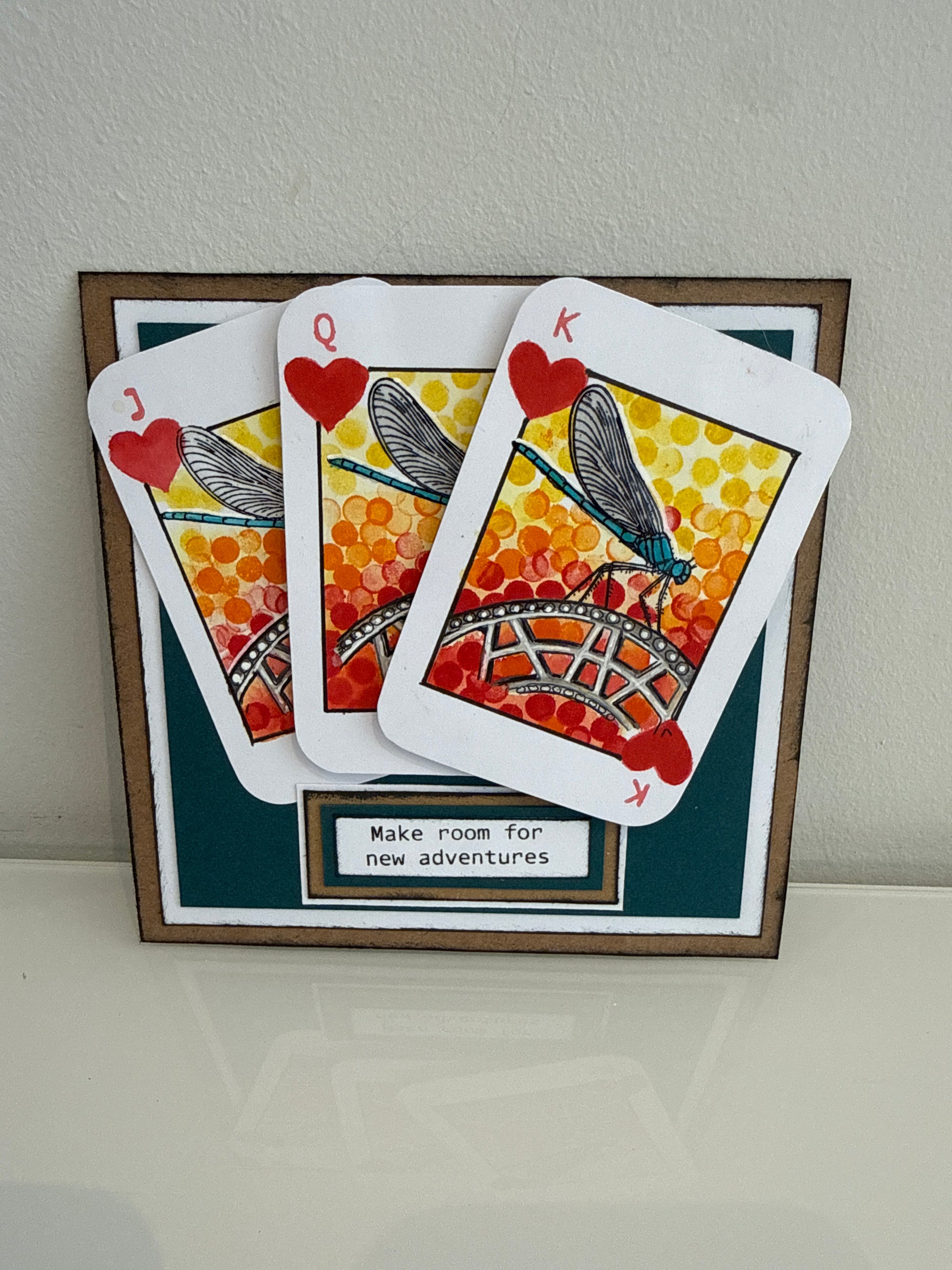 A framed decorative display featuring three overlapping playing cards styled like insect-themed cards with red hearts and orange-yellow patterns, mounted on a dark teal backing with a small label that reads 'Make room for new adventures.'.