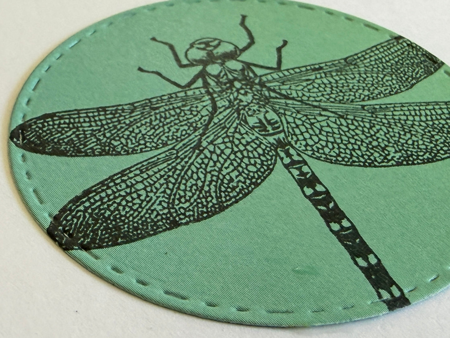 A round green coaster featuring a black printed dragonfly design and visible stitched edges, photographed on a light neutral background.
