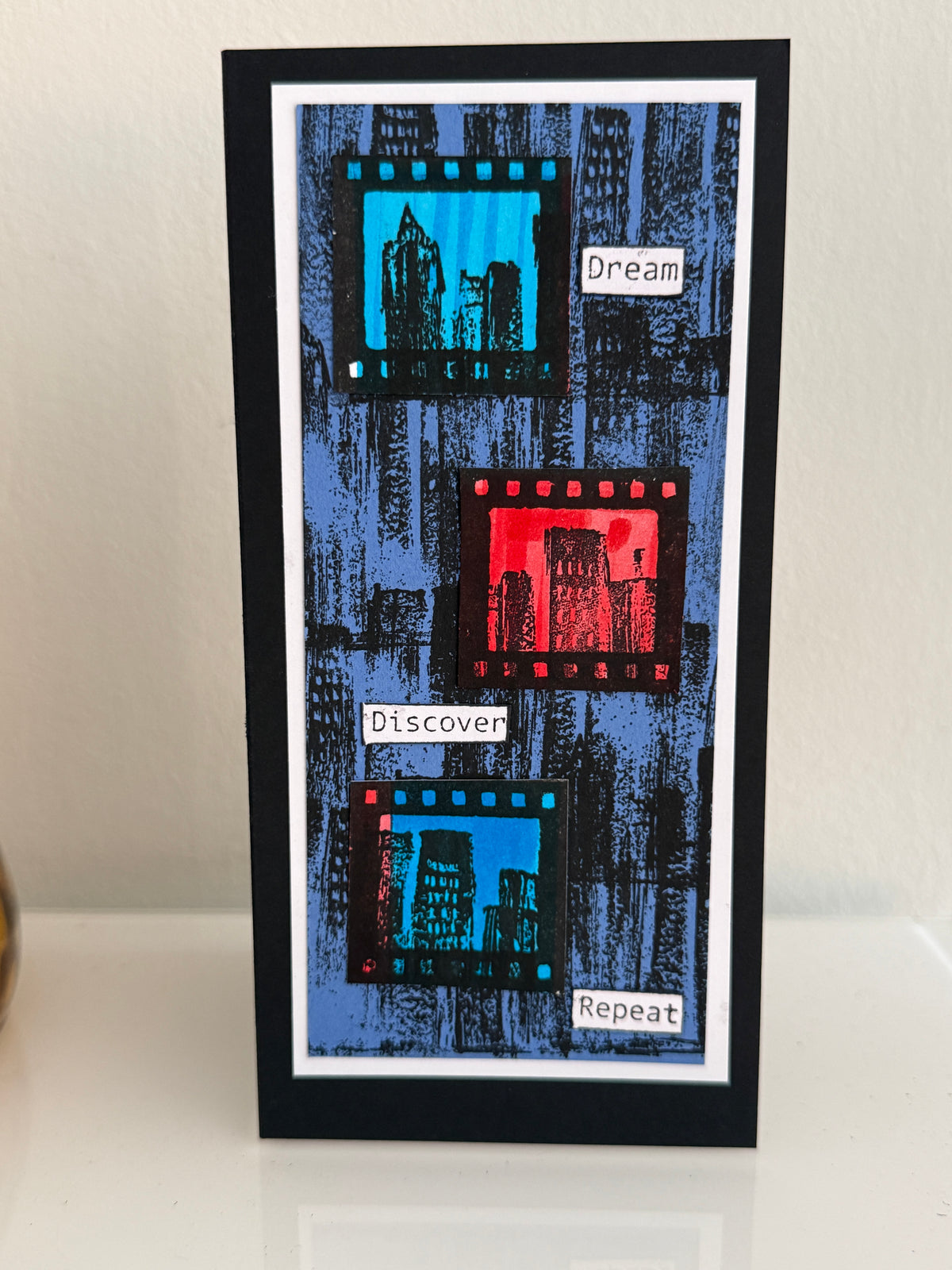 A handmade greeting card with a black border, featuring a blue textured background with three film strip graphics in black, red, and blue, and the words 'Dream', 'Discover', and 'Repeat' written on small white labels.