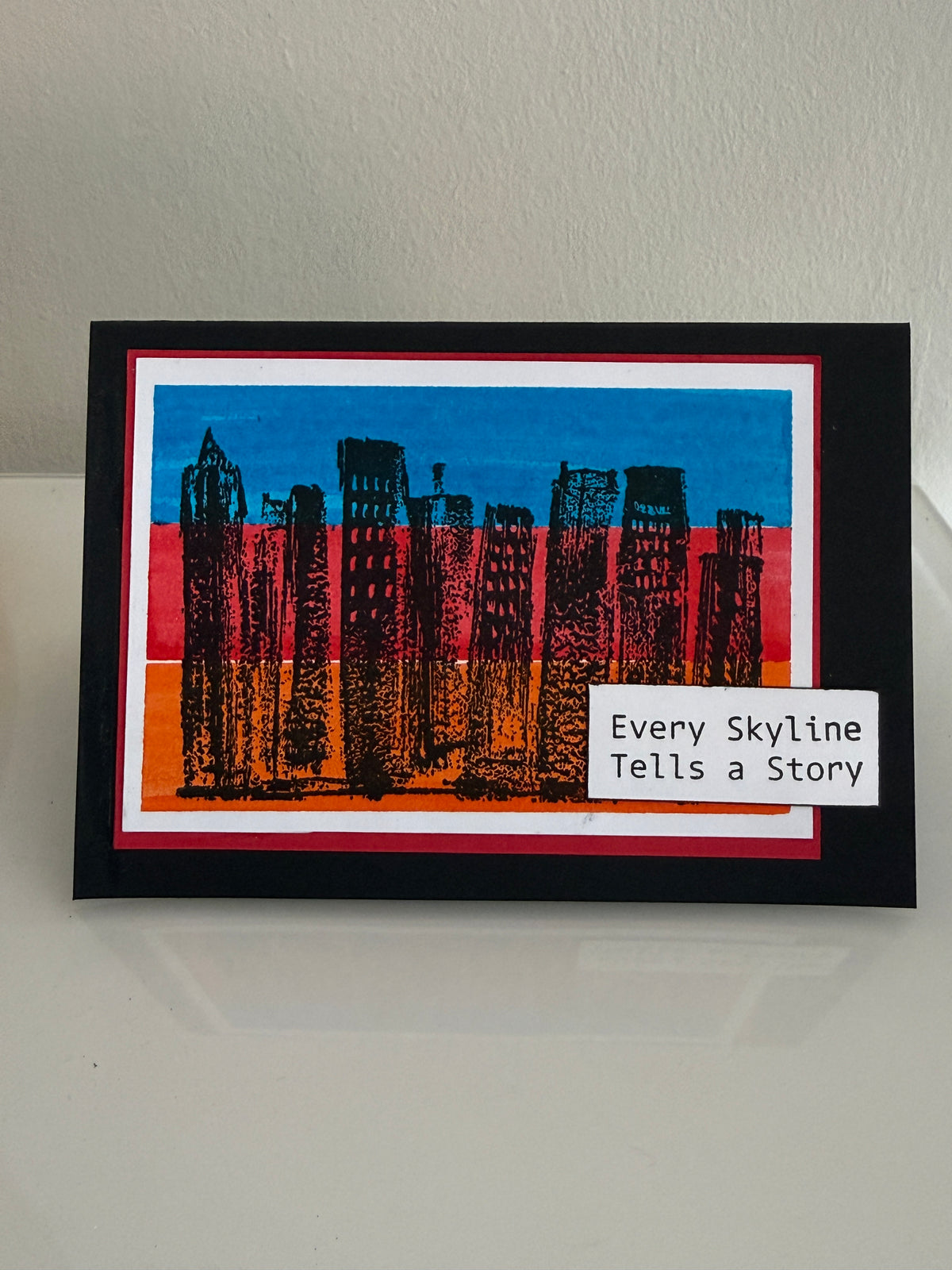 A square black-framed art print featuring a stylized city skyline in black ink over blue, red, and orange blocks, with a white label reading 'Every Skyline Tells a Story' on the lower right, photographed against a light wall.
