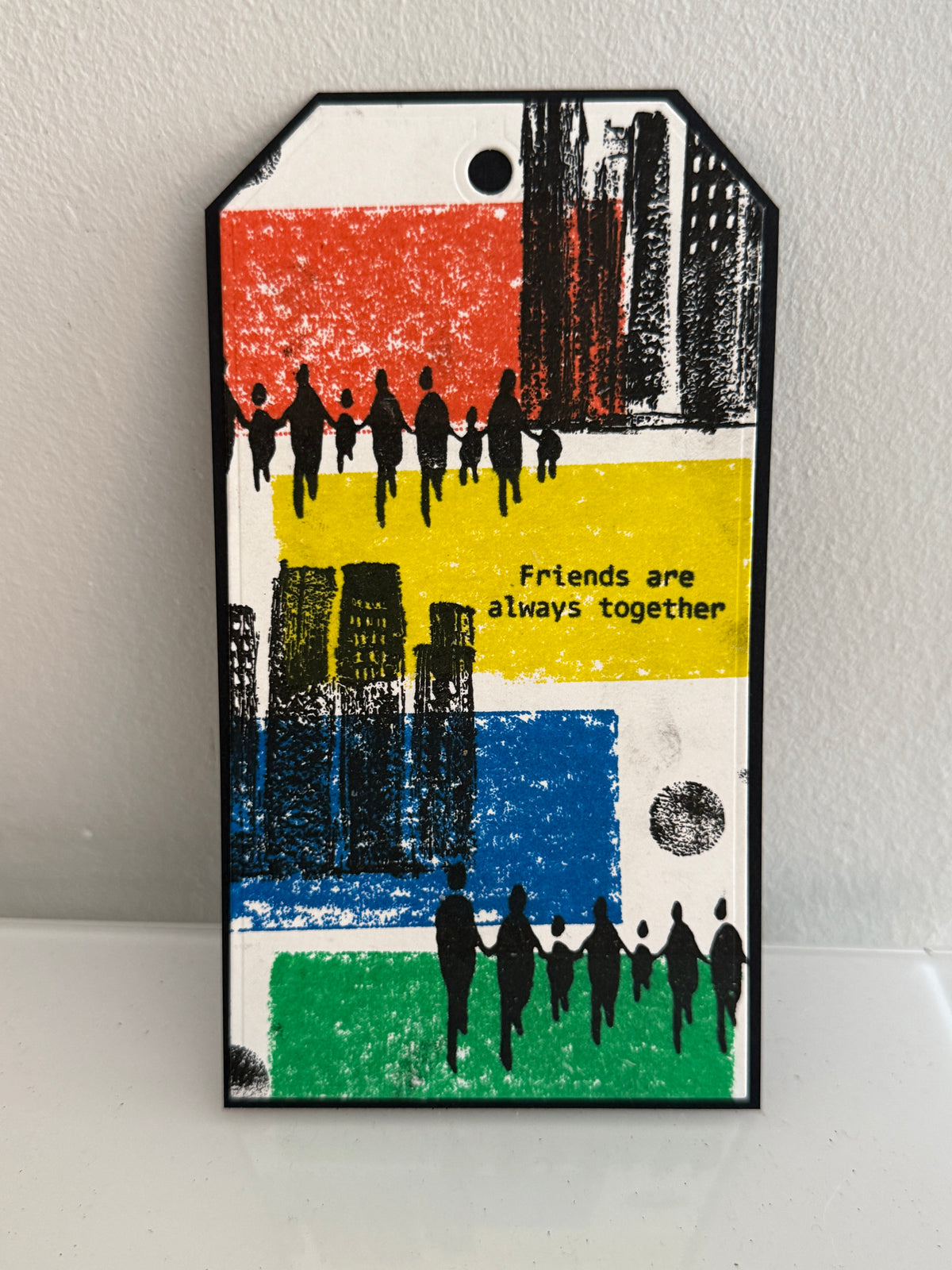 A rectangular decorative tag with a black border and a small hanging hole, featuring a multicolor abstract cityscape design with black silhouettes of people and the text 'Friends are always together' on a white background.