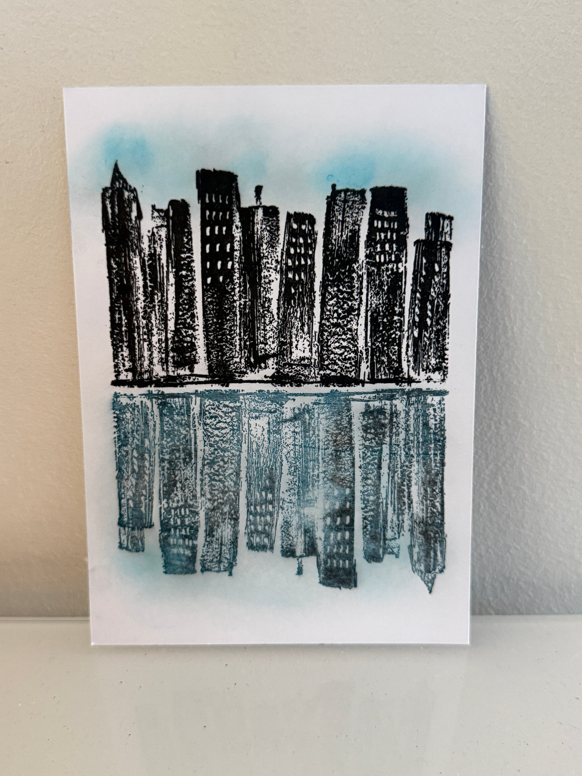 A vertical art print featuring a black ink city skyline with reflected buildings and water, printed on white paper and leaning against a light beige wall.