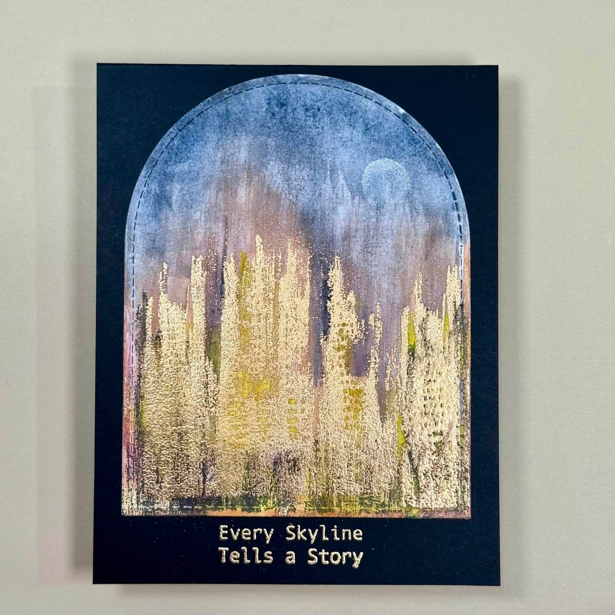 A square art print or card featuring an abstract skyline scene under a blue sky with a pale moon, bordered by a dark navy frame with the text 'Every Skyline Tells a Story' at the bottom, photographed against a light neutral wall.