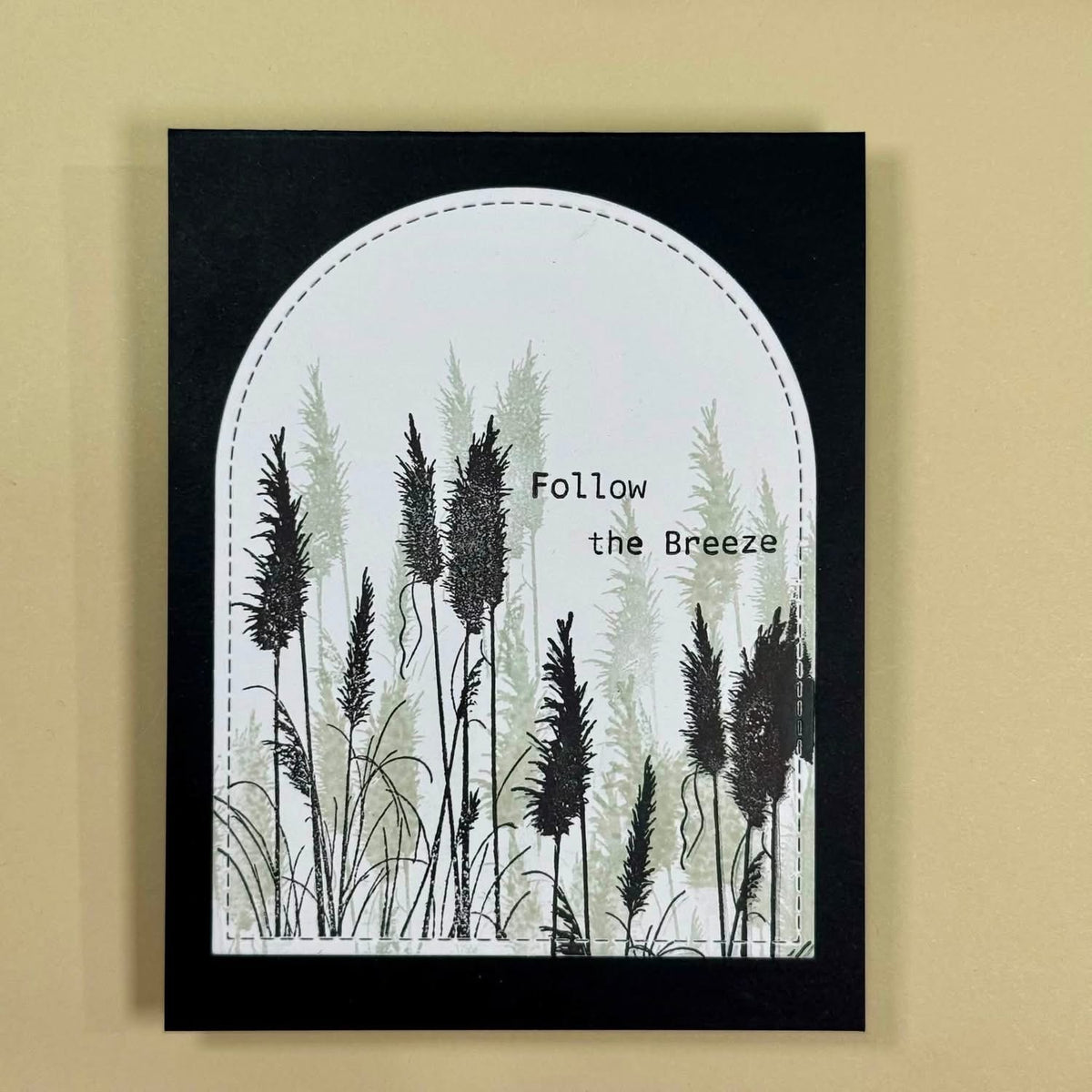 A handmade greeting card featuring a black arched border with a white center, decorated with black and muted green reeds and the text 'Follow the Breeze'. The card is displayed flat against a warm beige background.