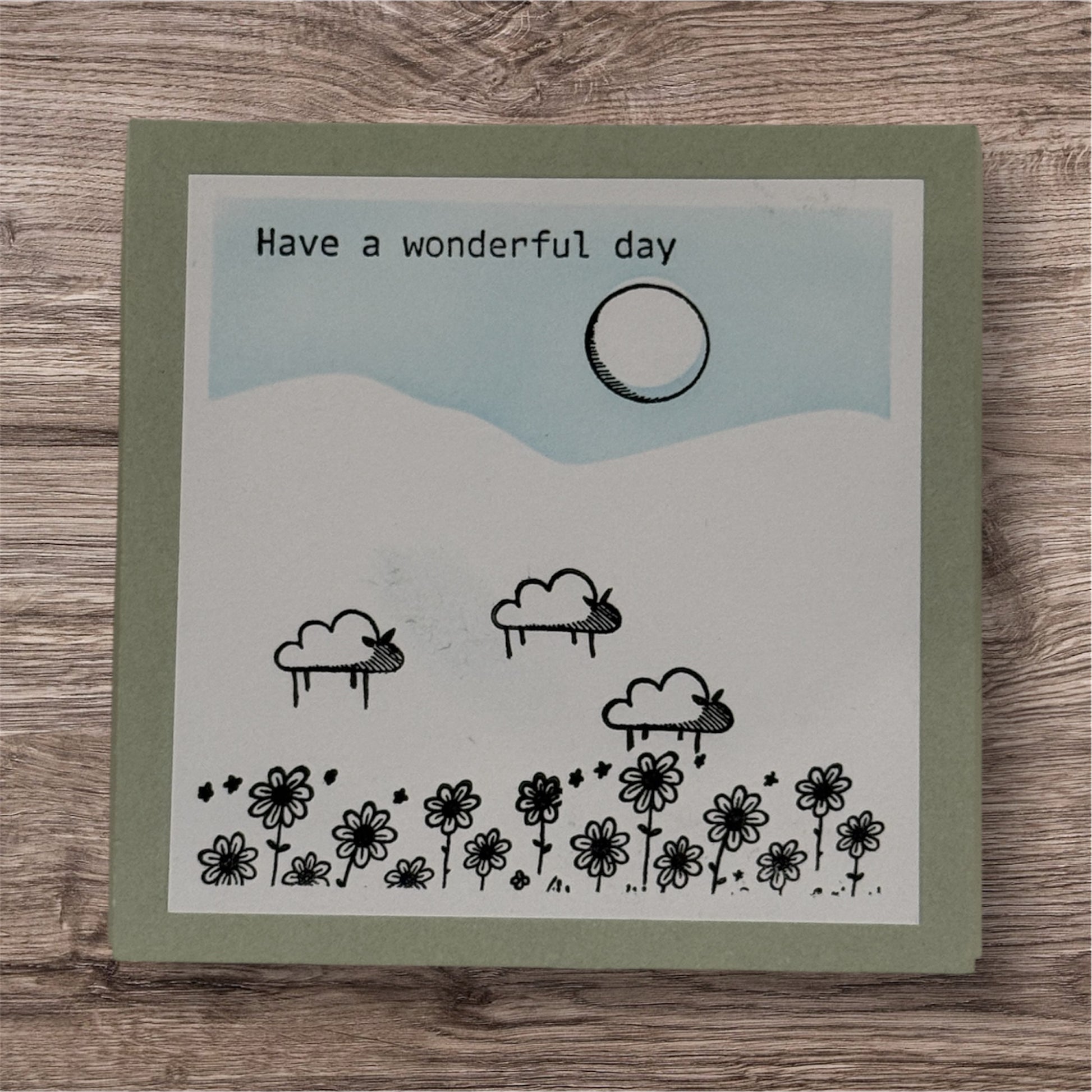 Card with a hand-drawn landscape, flowers, and clouds with 'Have a wonderful day' text on a wooden surface.