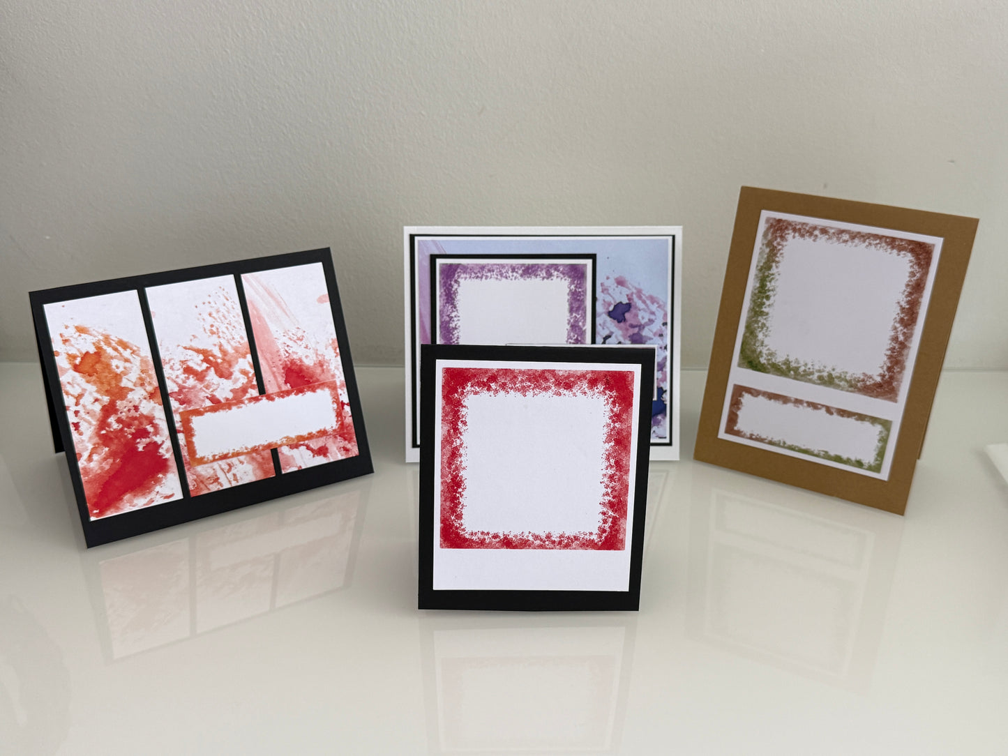 Three handmade greeting cards with textured stamp borders are displayed on a glossy white surface against a light beige wall. One card features red and orange splatters on a black background, another has a red textured border, and the third shows a green textured border on a kraft brown card.