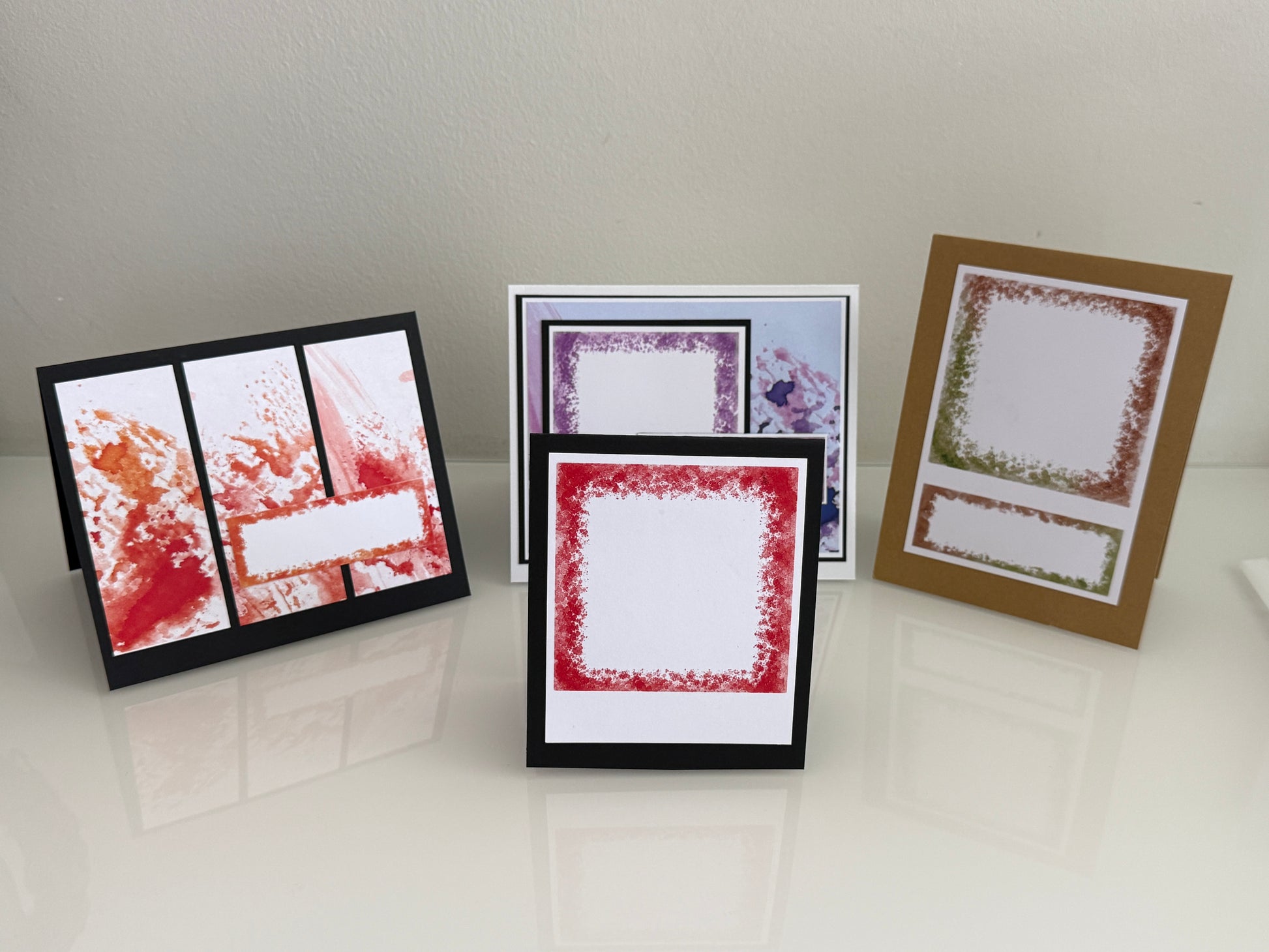 Three handmade greeting cards with textured stamp borders are displayed on a glossy white surface against a light beige wall. One card features red and orange splatters on a black background, another has a red textured border, and the third shows a green textured border on a kraft brown card.