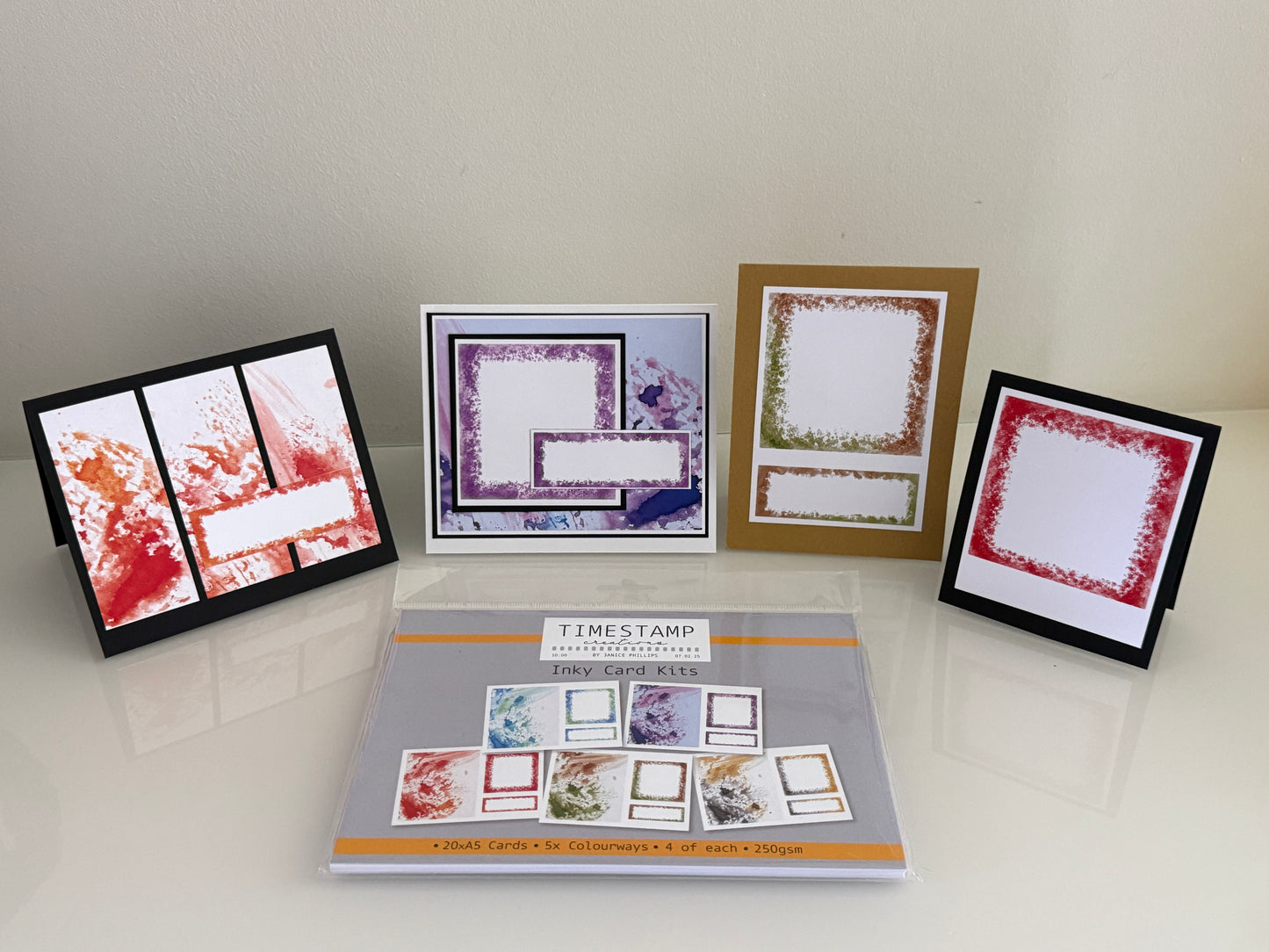 A collection of handmade greeting cards and a stamp booklet displayed on a glossy white surface against a beige wall. The set includes three framed cards with red, purple, and green stamp designs, and a central stamp booklet labeled 'TIMESTAMP QUOTED CARD SET' by Stampin' Up!.