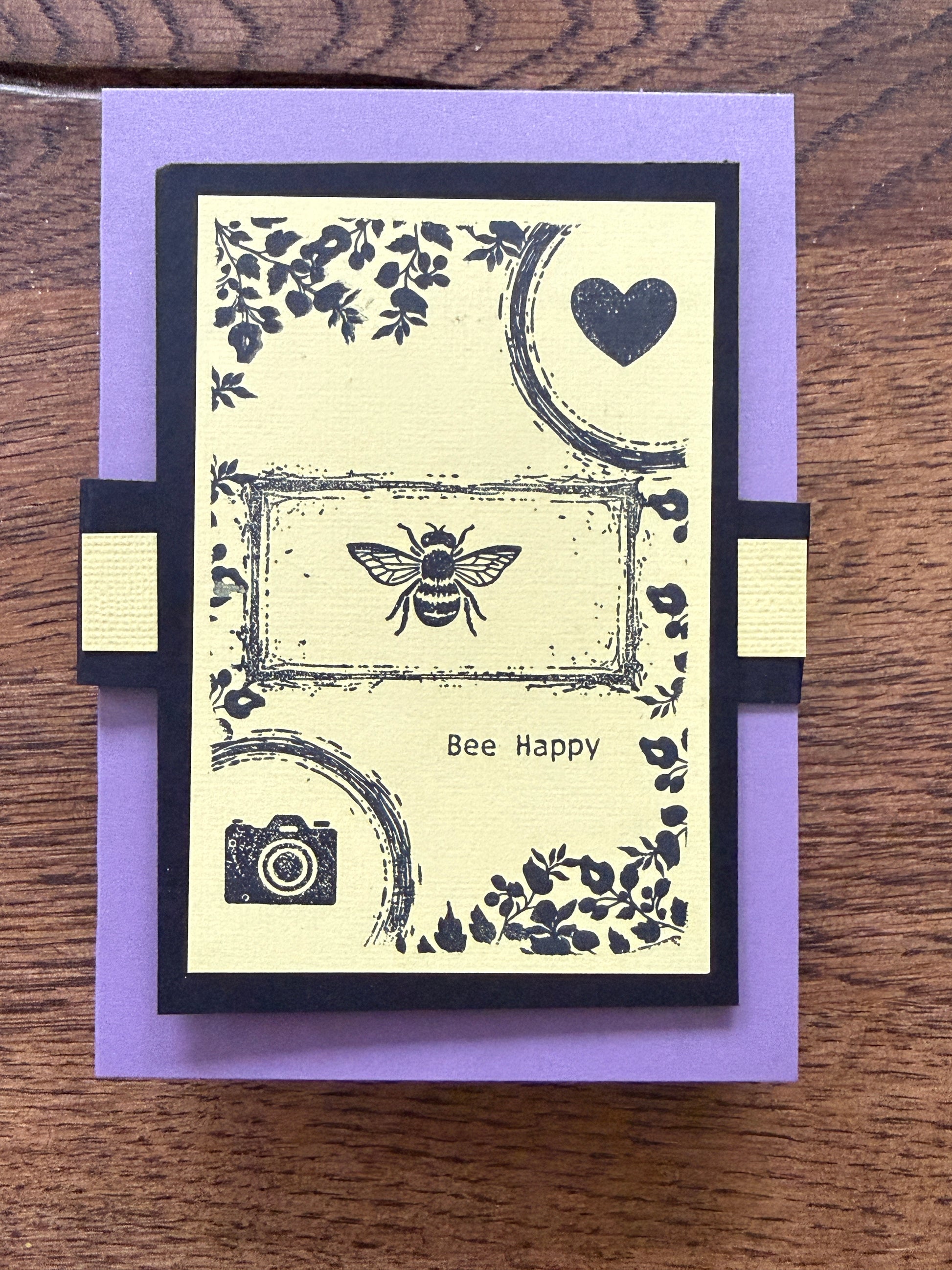 A handmade greeting card featuring a black and cream design with a bee, heart, camera, and floral motifs, displayed on a purple cardstock base over a wooden surface.